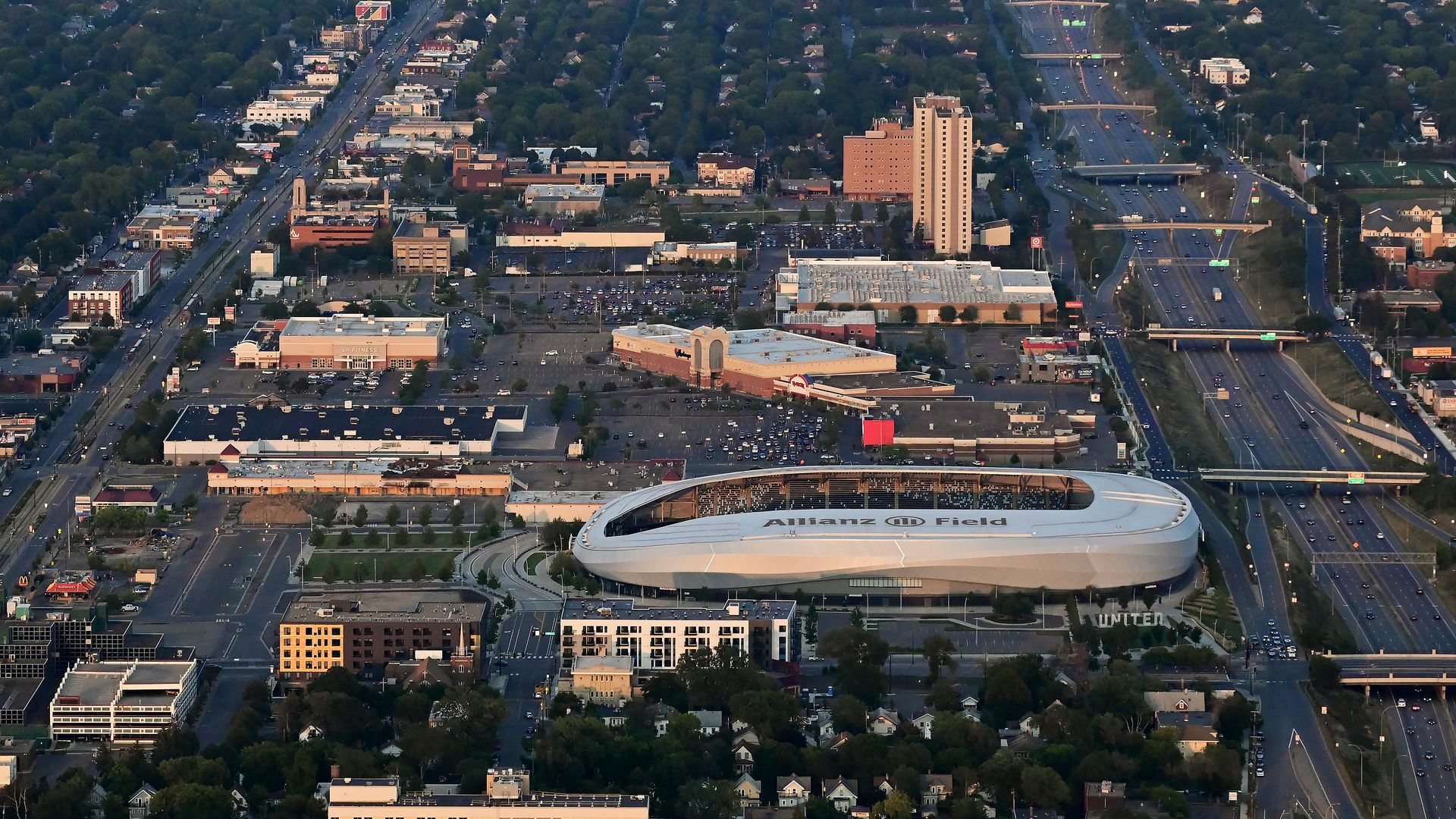 Aerial view of a dense city with green suburbs, a long central avenue, and a distant skyline; a large white oval stadium labeled Allianz Field sits in the foreground.