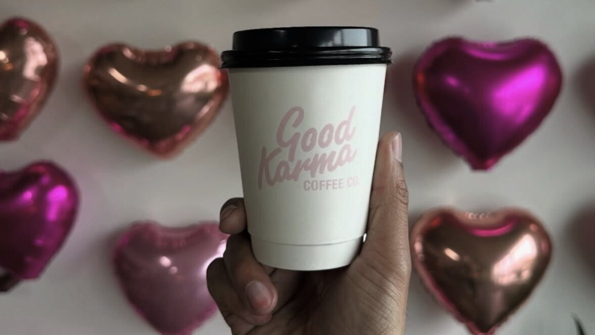 Hand holding a white coffee cup with black lid from Good Karma Coffee Co., with metallic pink and rose gold heart-shaped balloons blurred in the background.