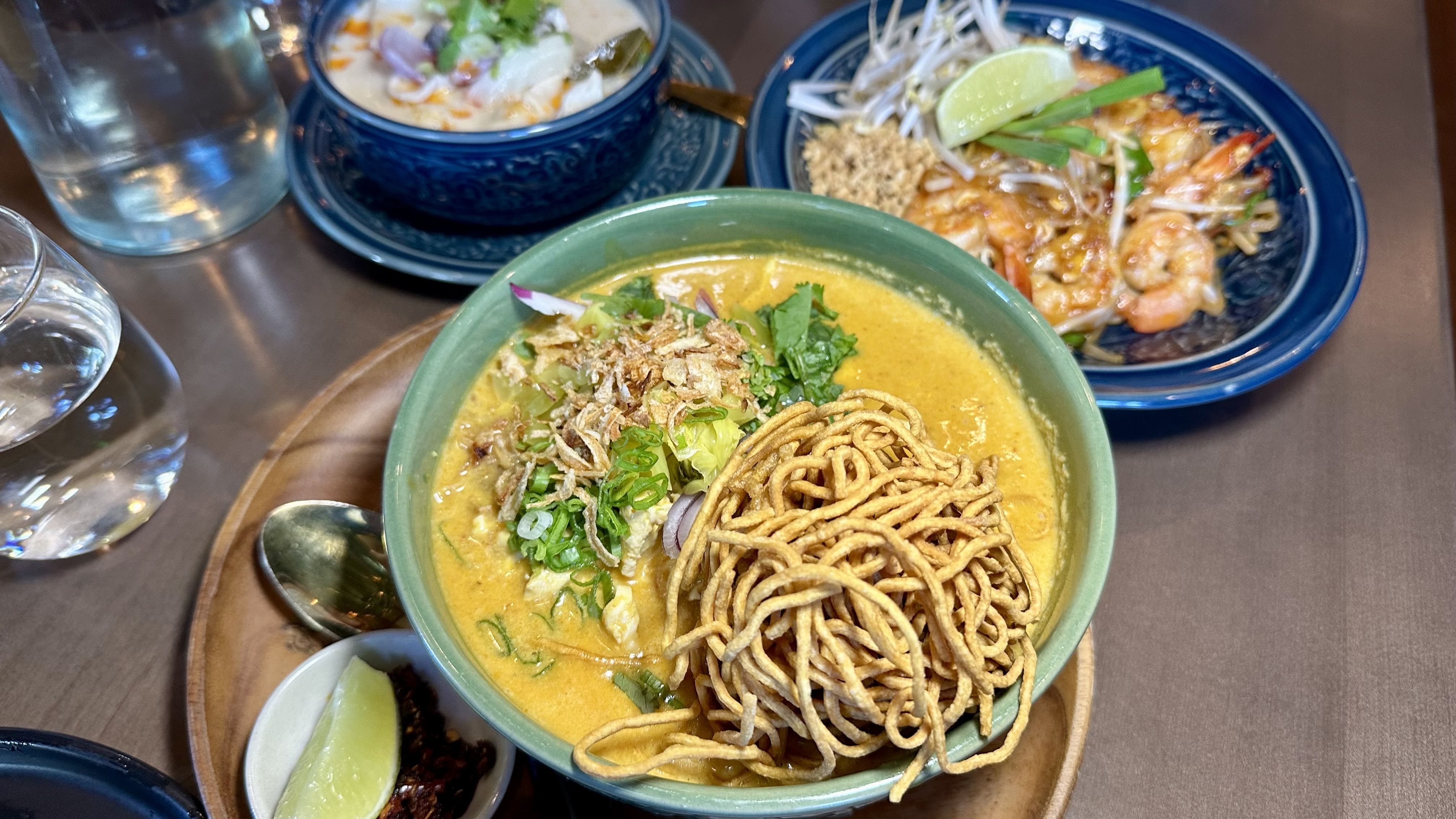 Top-down view of a table with a green bowl of curry laksa topped with crispy noodles, scallions, and cilantro; blue plates hold shrimp, bean sprouts, lime, and a side creamy soup, plus a glass of water.