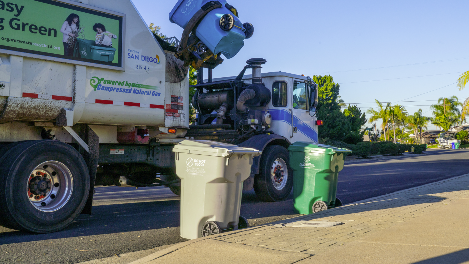 A garbage truck with a mechanical arm lifts a light blue bin on a street. Two wheeled trash cans, gray and green, sit on the curb beside the sidewalk, with palm trees and houses in the background.