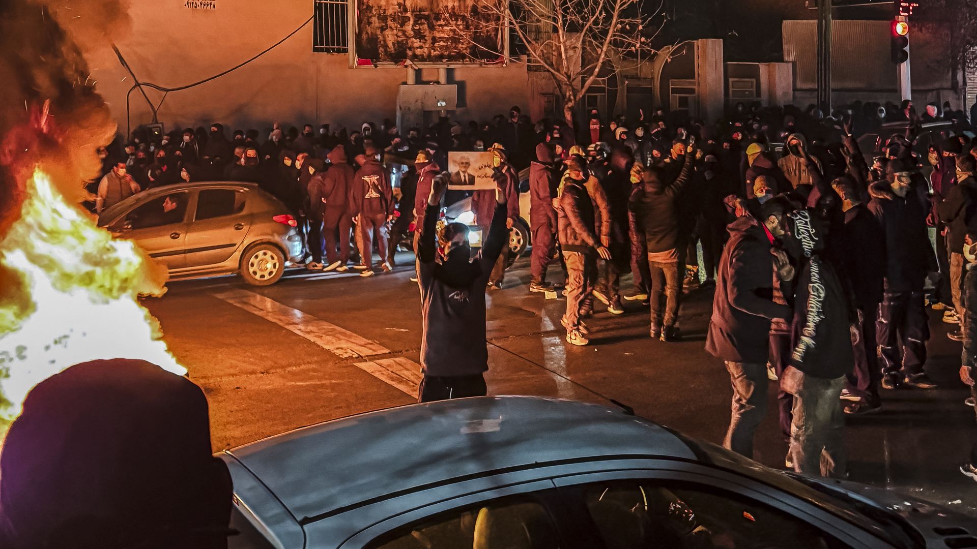 Dozens of protesters are seen to the right of the photo. In the center, one protester wearing a mask holds up a sign. On the left, there is a fire. 