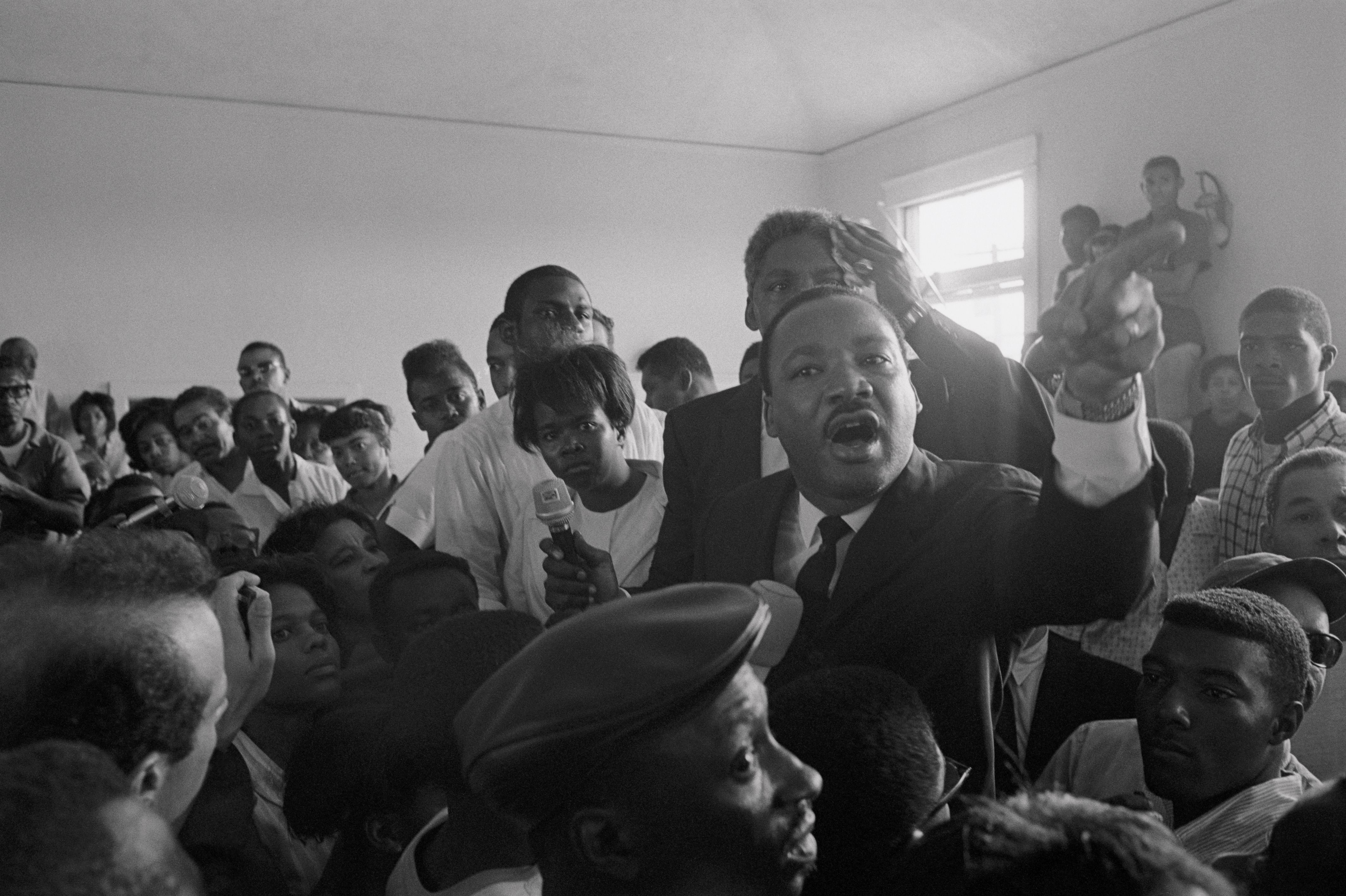 Black and white photo showing Martin Luther King Jr. speaking passionately to a crowd indoors, surrounded by attentive people, some holding microphones and cameras.