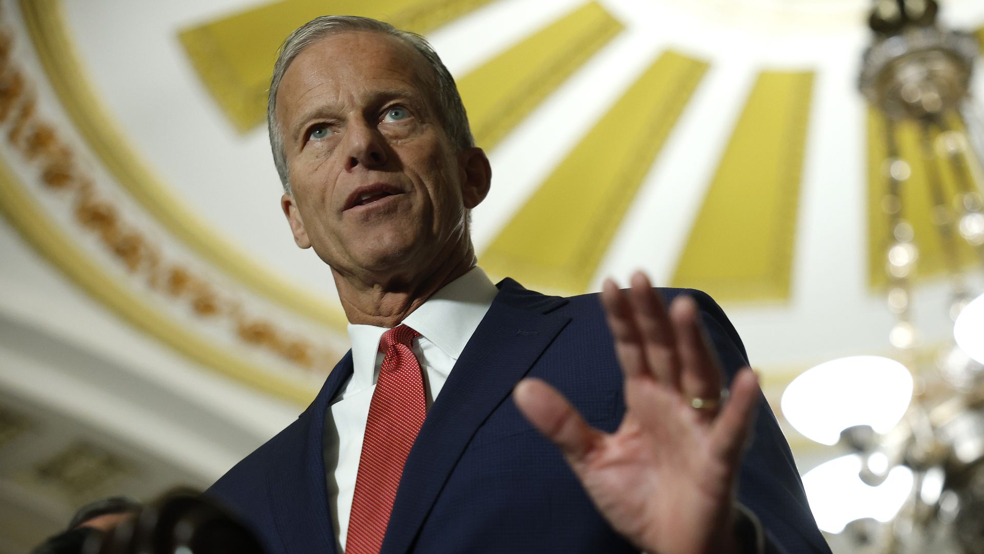 John Thune, wearing a dark suit and a red tie with a white shirt, holds his hand up as he speaks on Capitol Hill. The gold roof and chandelier are visible behind him. 