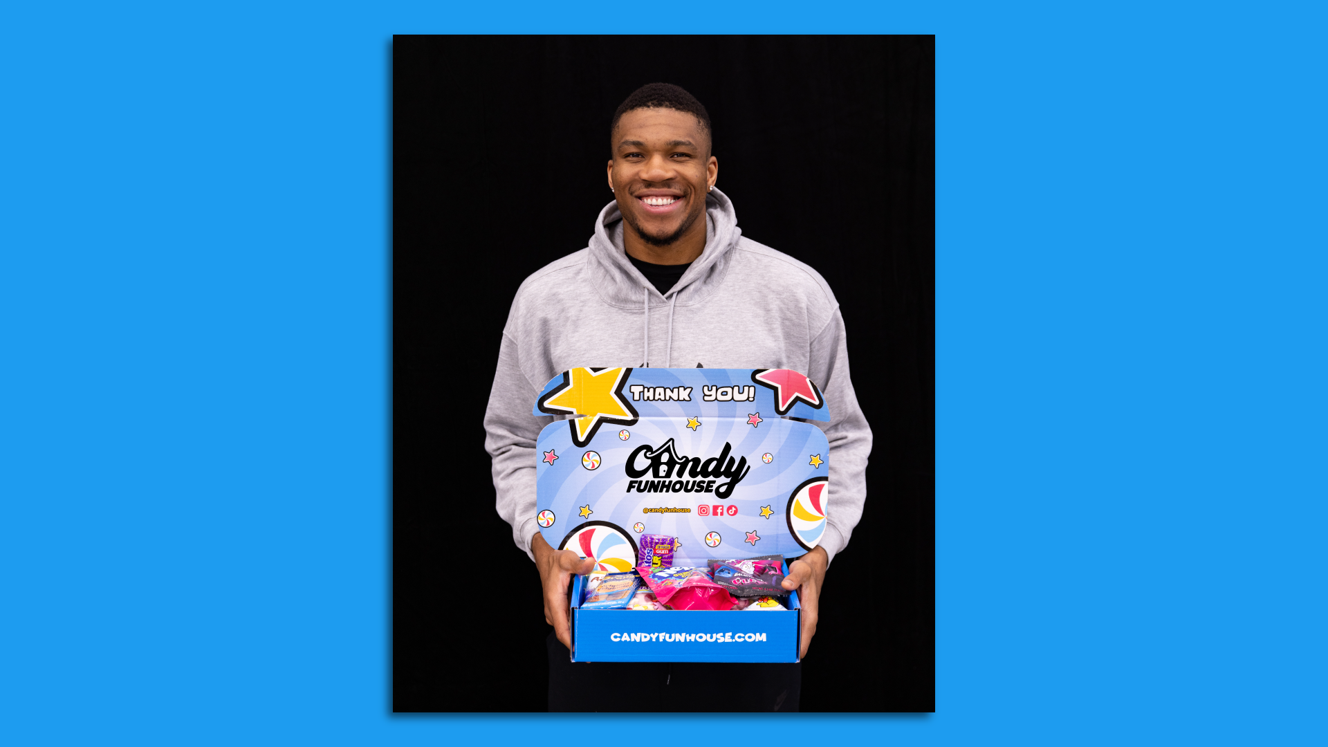 Giannis Antetokounmpo standing holding a blue box from Candy Funhouse that is filled with assorted candies