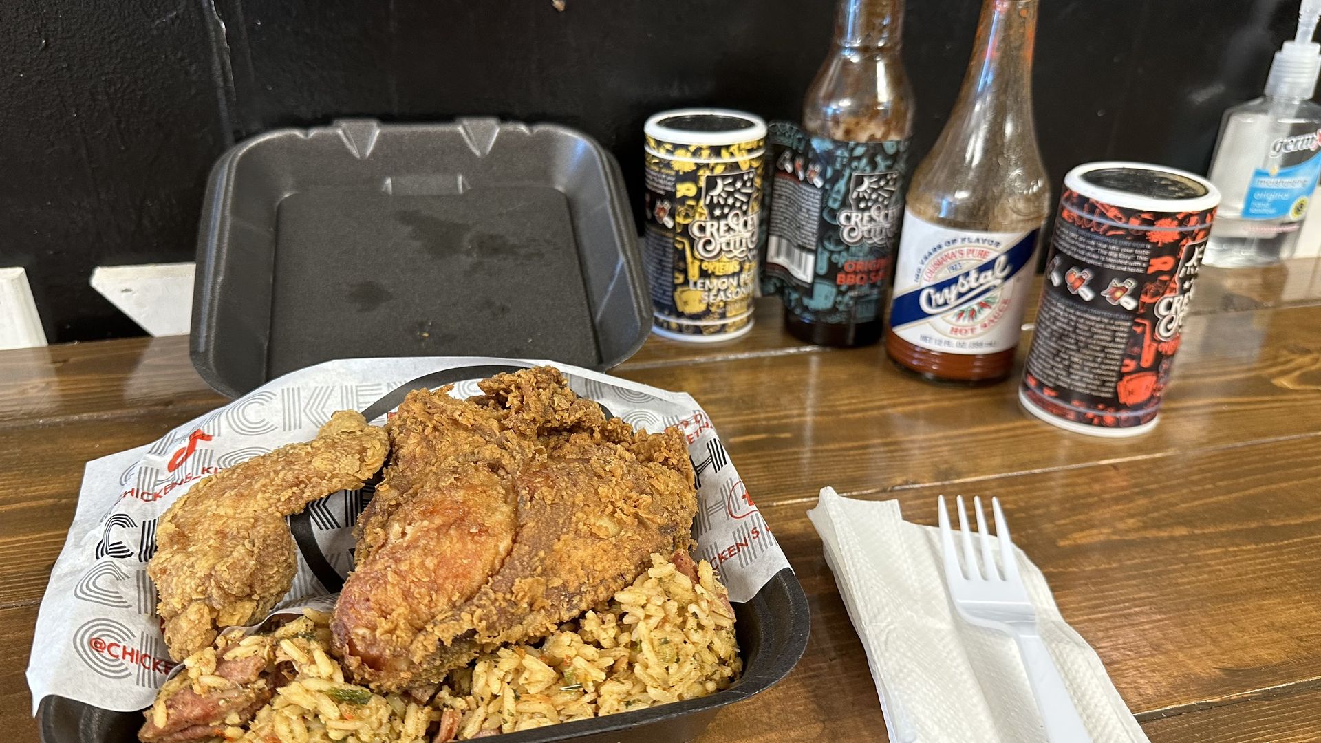 Fried chicken sits atop an open and very full styrofoam clamshell takeout container. Jambalaya is seen beneath the chicken, and hot sauces are set to the side of the container.