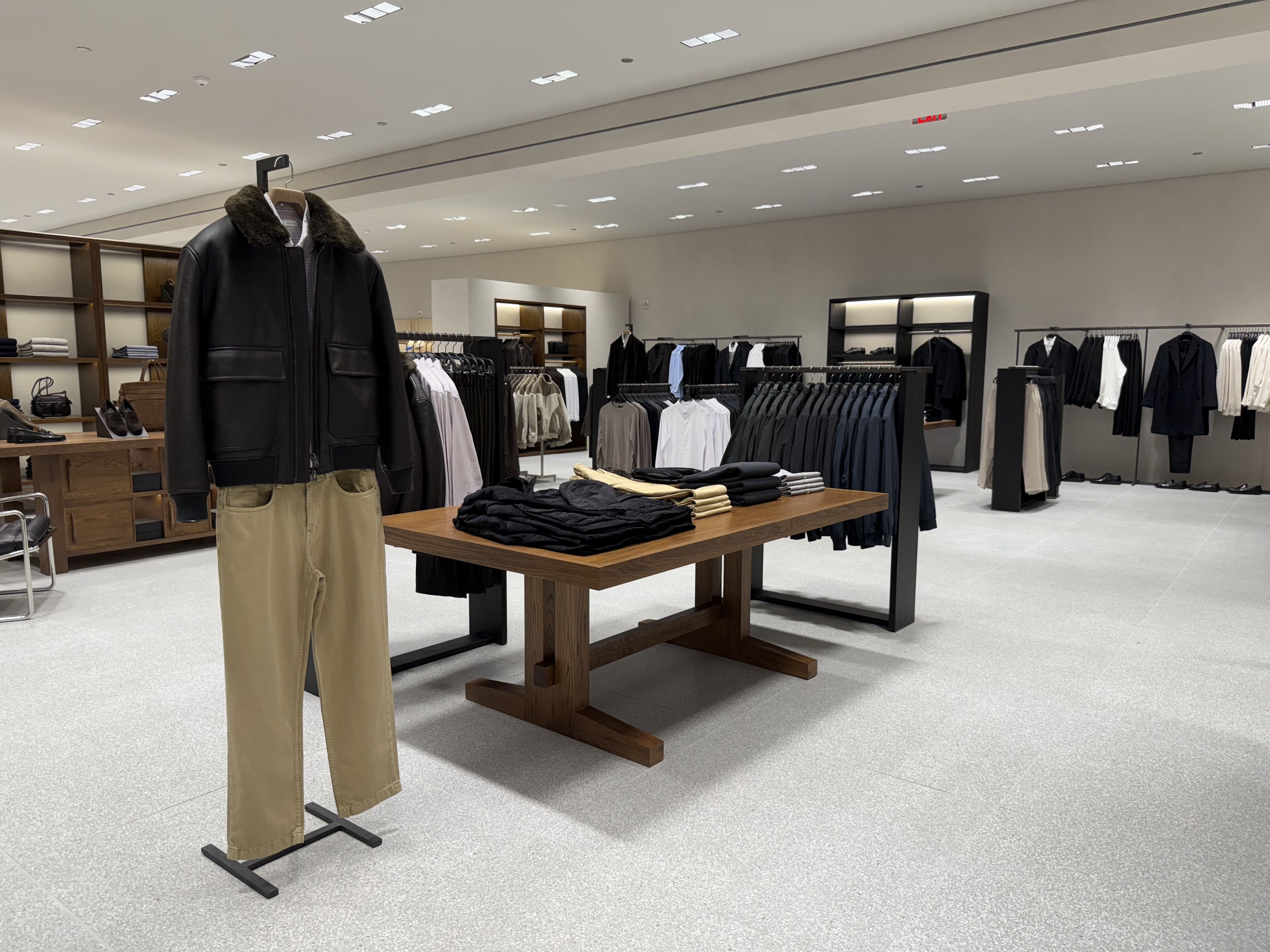 Modern clothing store interior with beige pants and black leather jacket on display, wooden table with folded black and beige clothes, racks of neatly arranged shirts and pants under bright lights.