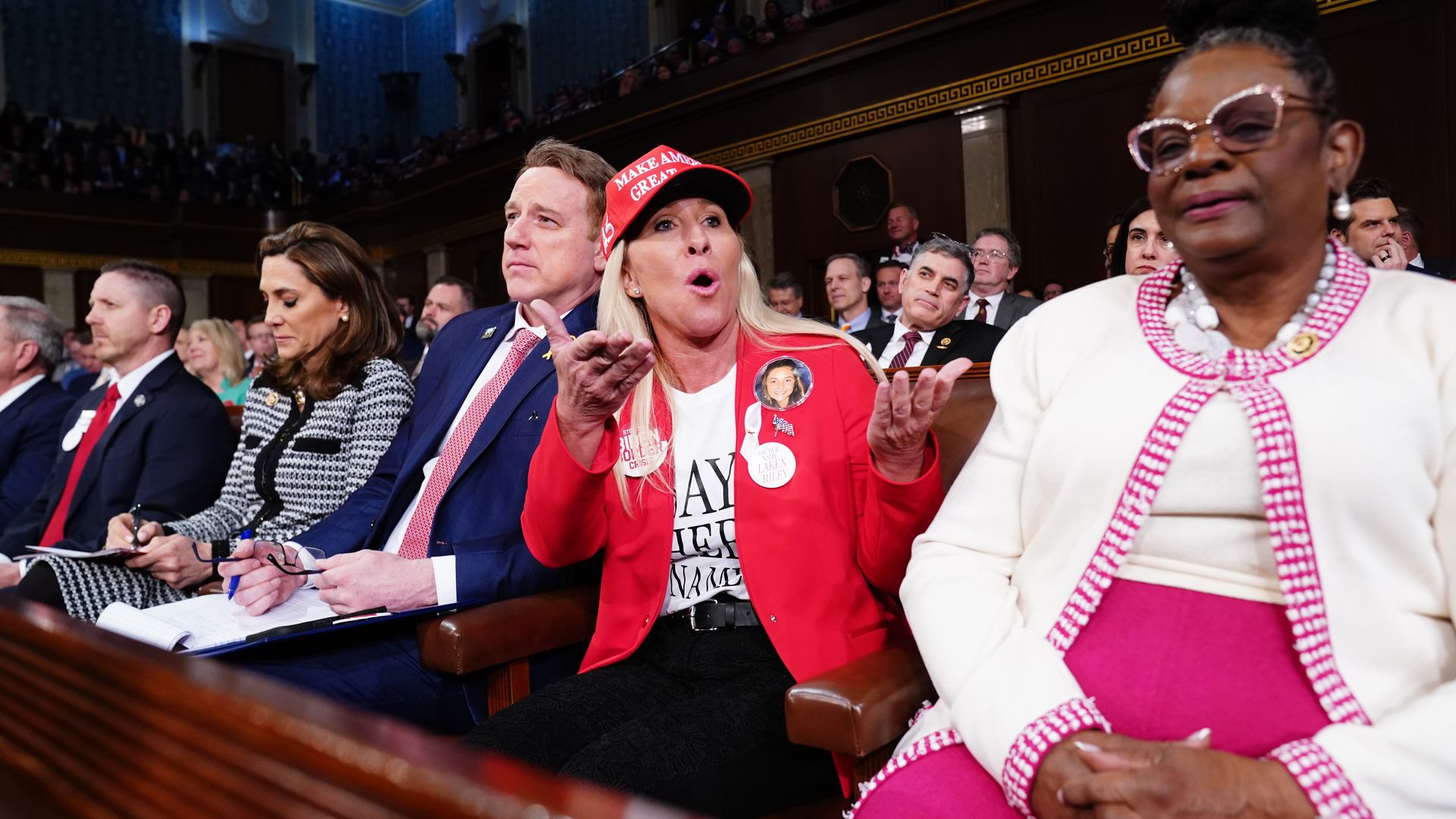 Rep. Marjorie Taylor Greene, wearing a red blazer, white shirt, buttons and red hat, seated with colleagues in the House chamber.