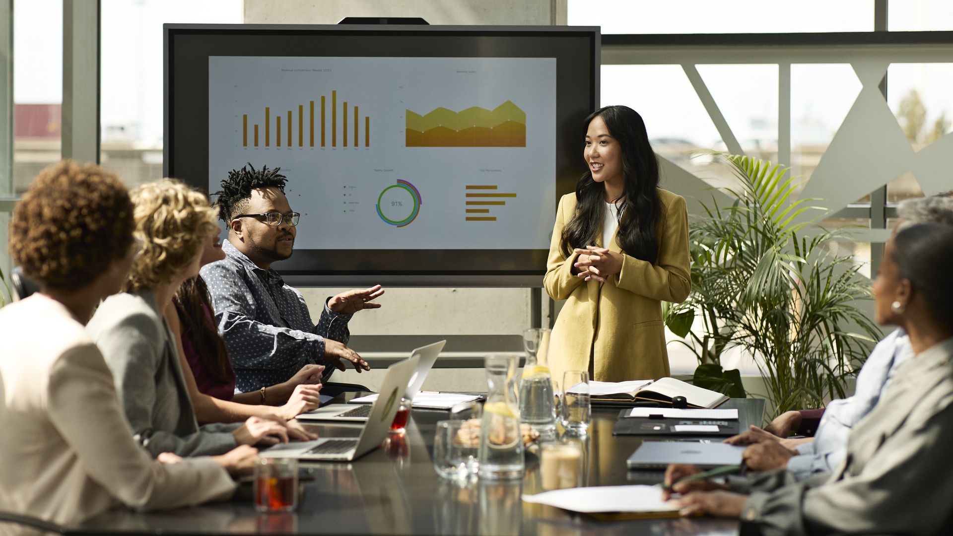 A woman in a yellow blazer presents business charts on a screen to a diverse group of colleagues seated around a conference table with laptops and notebooks in a bright office.