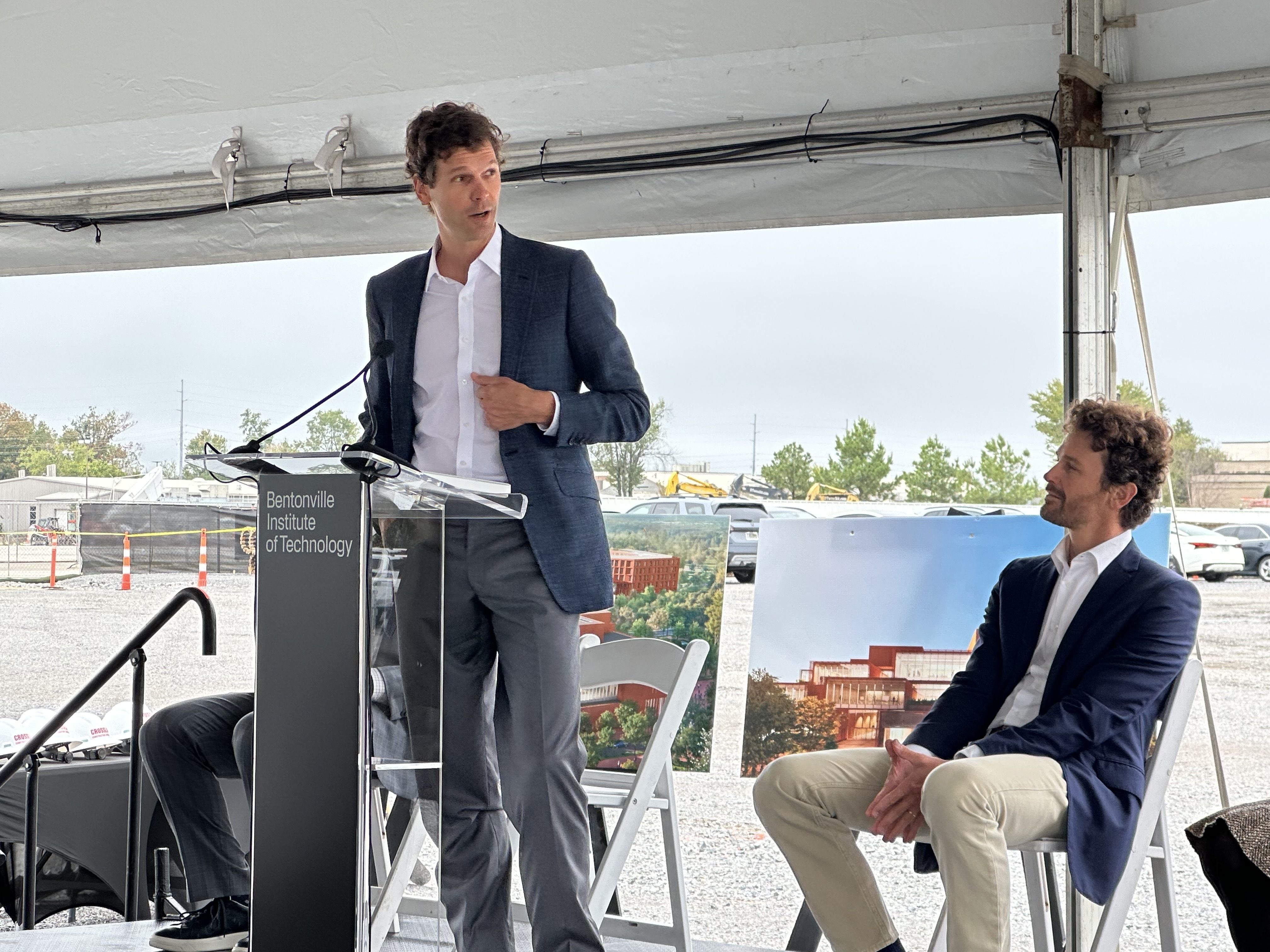 Two men in suits under a white tent; one speaks at a lectern labeled "Bentonville Institute of Technology," the other sits nearby as a construction site and parked cars appear in the background.