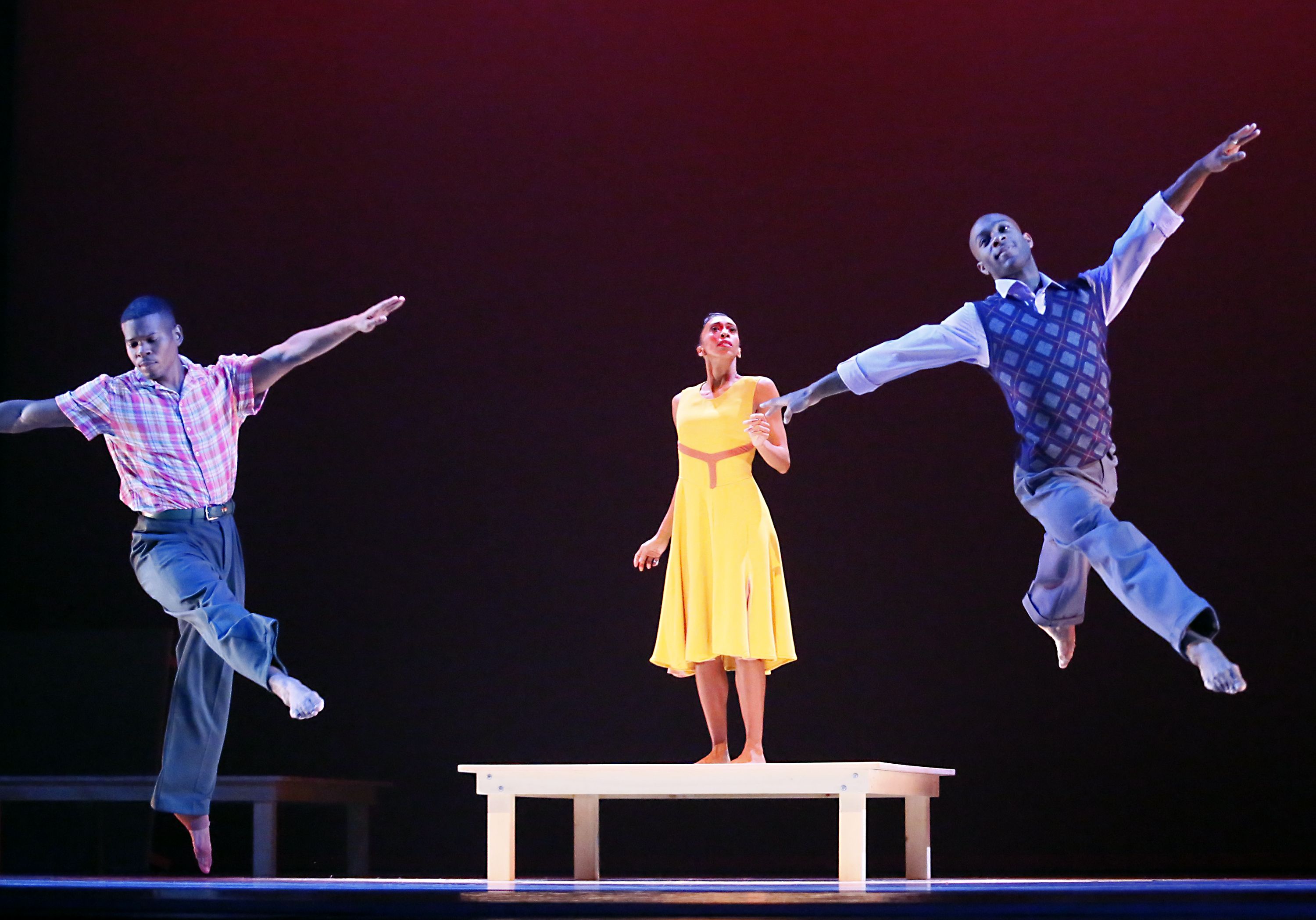 Three Alvin Ailey dancers on stage with a dark red background: a woman in a yellow dress stands on a white platform while two men in pants and shirts leap on either side of her.