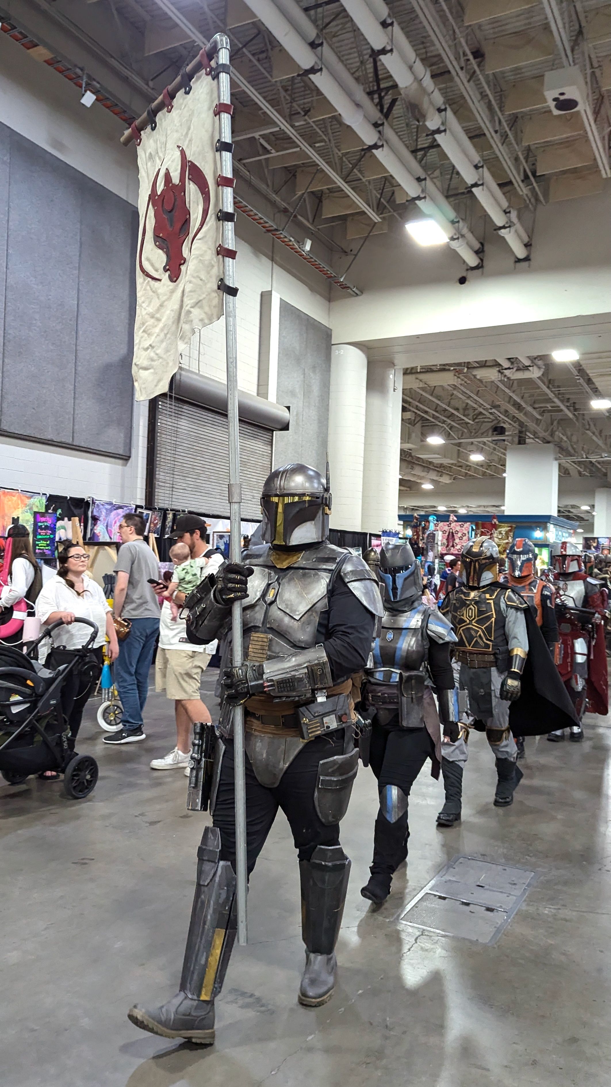 A parade of people costumed as Mandalorians from Star Wars march through a convention center behind a banner with a Mandalorian crest