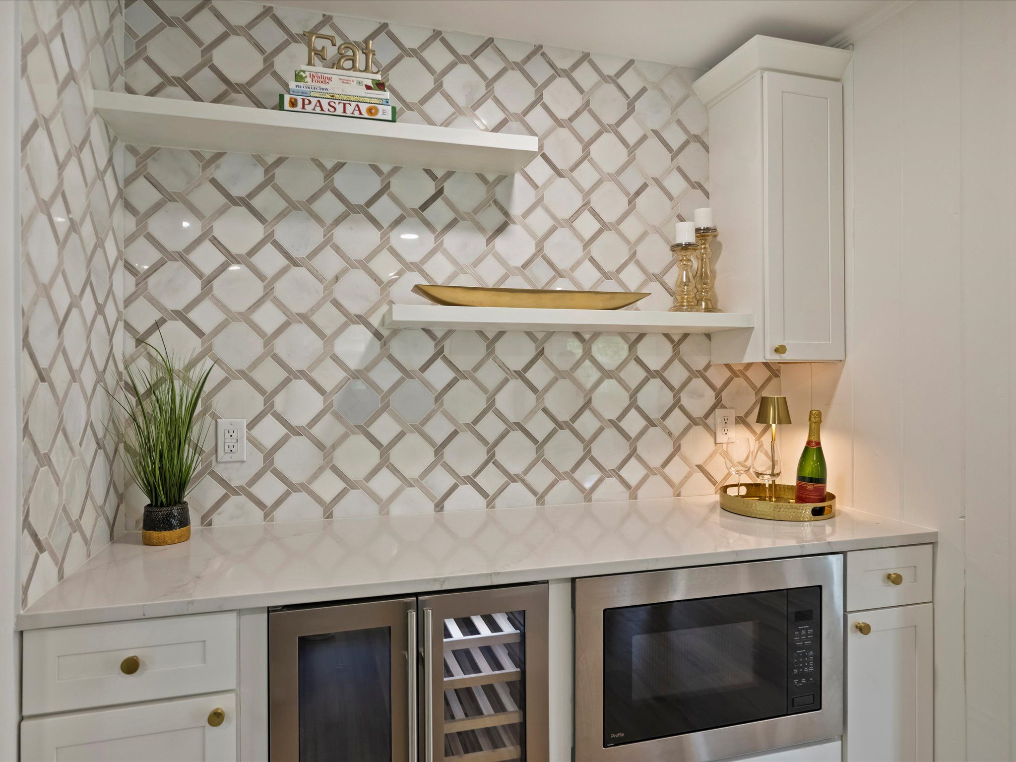 Modern kitchen corner with patterned beige and white tile backsplash, white shelves holding books, a gold tray, candle holders, and a small green plant pot. Stainless steel wine fridge and microwave below.