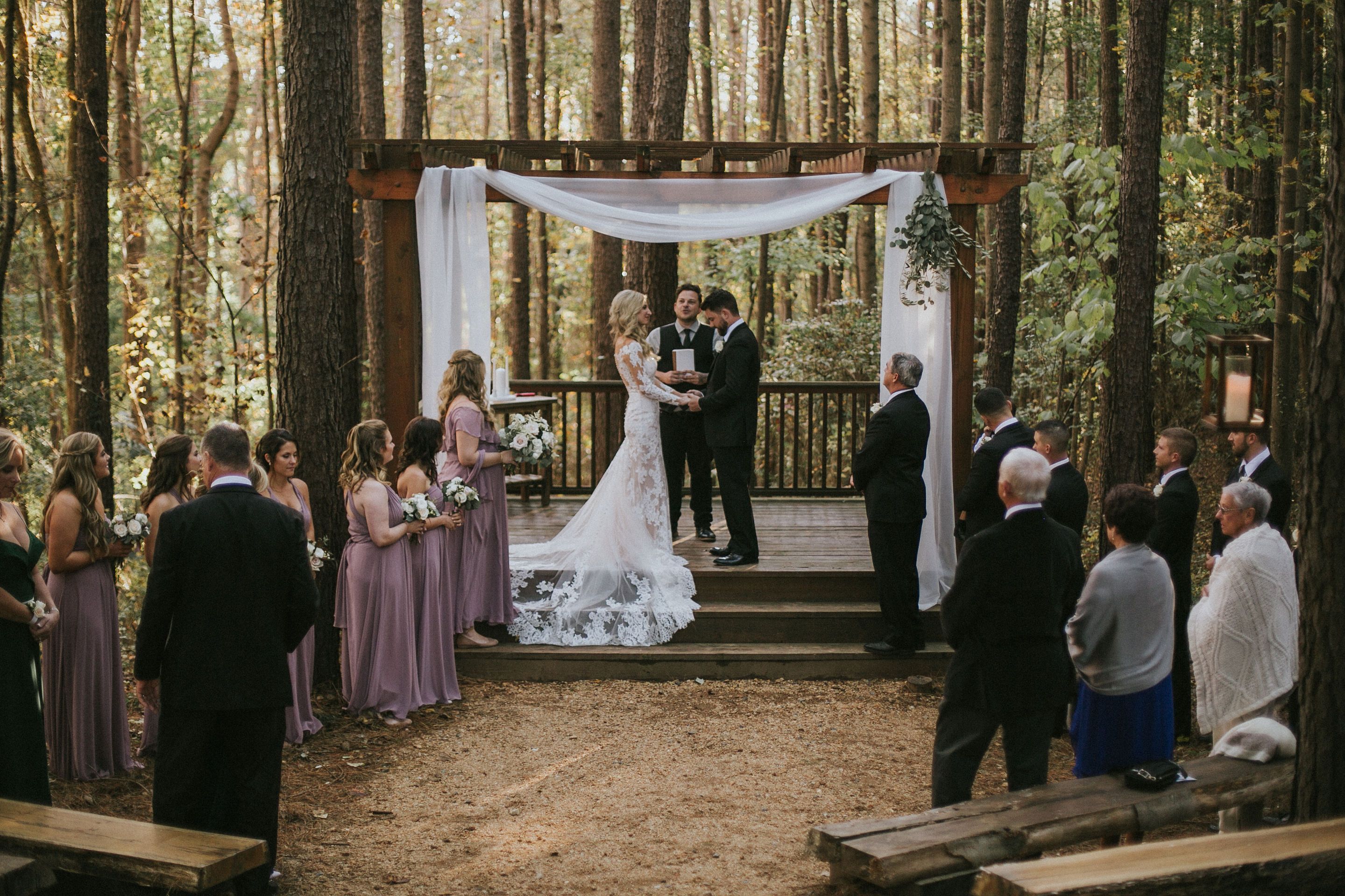 Outdoor wedding in a forest setting with bride in white lace gown and groom in black suit holding hands under a wooden arch draped with white fabric, surrounded by bridal party and guests at the Whitewater Center. 