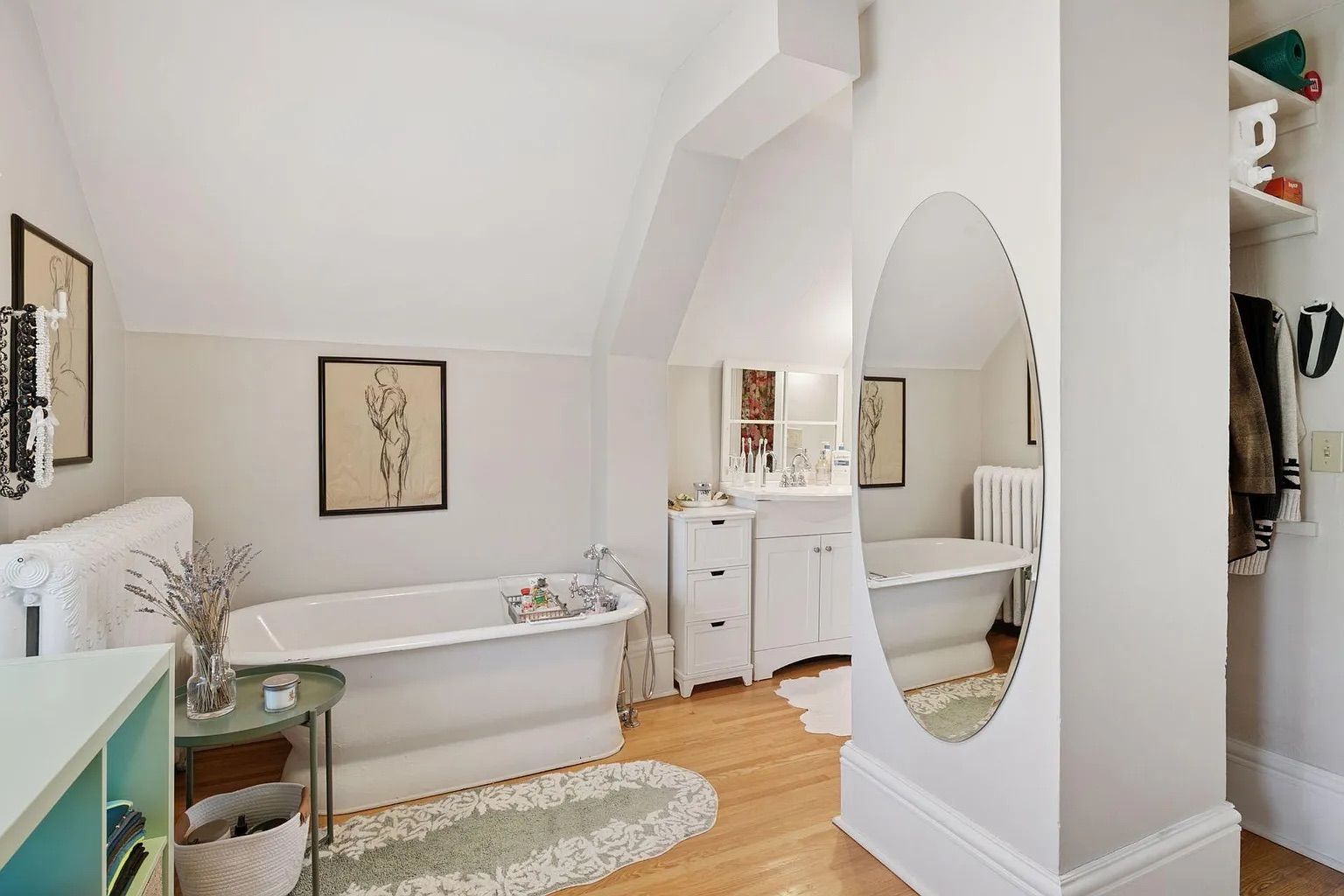 Bright bathroom with white freestanding tub, light wood floor, oval wall mirror, white cabinets, framed art, small green table with decor, and open closet with clothes.