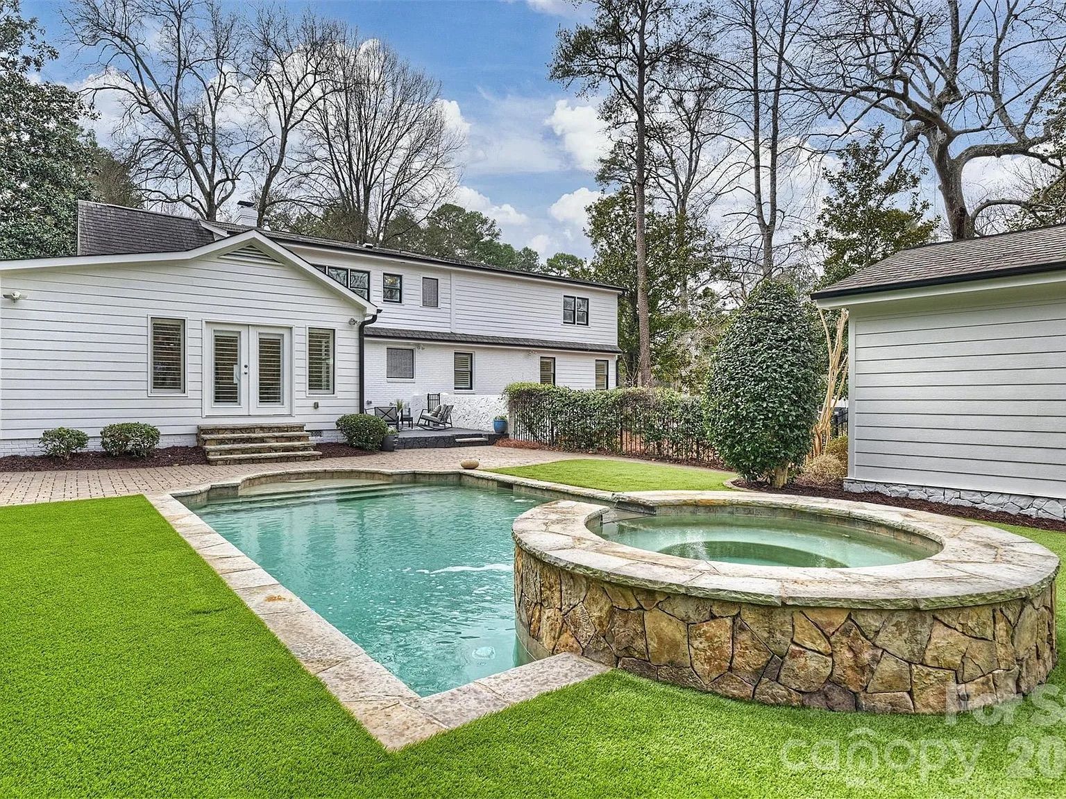 Backyard of a white two-story house with a rectangular pool and attached round stone hot tub, surrounded by green grass and leafless trees under a partly cloudy blue sky.