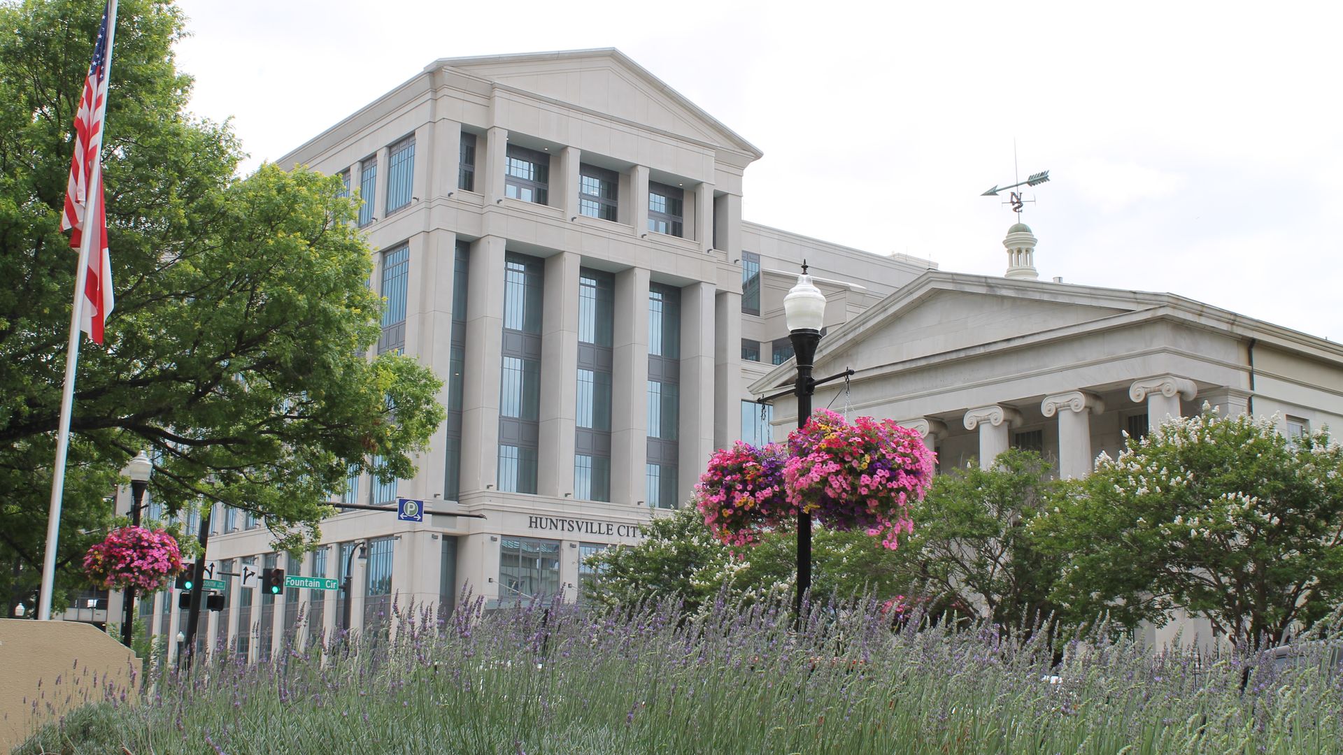 Huntsville City Hall building with columns, a weather vane on the roof, hanging baskets of pink flowers, green trees, and an American flag on the left under a cloudy sky.