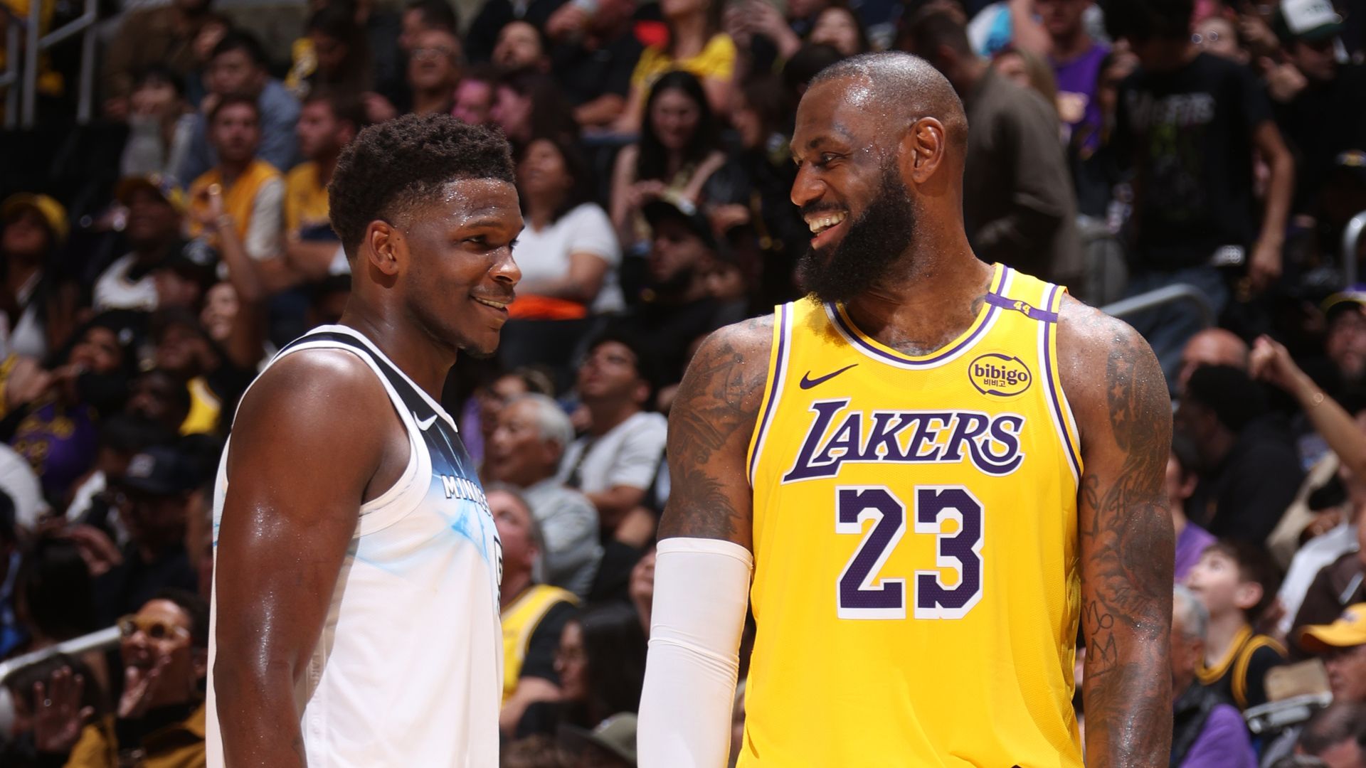 Two basketball players on a court smile at each other. Anthony Edwards in a Minnesota Timberwolves jersey and LeBron James in a Los Angeles Lakers jersey with number 23; a crowded arena is behind them.