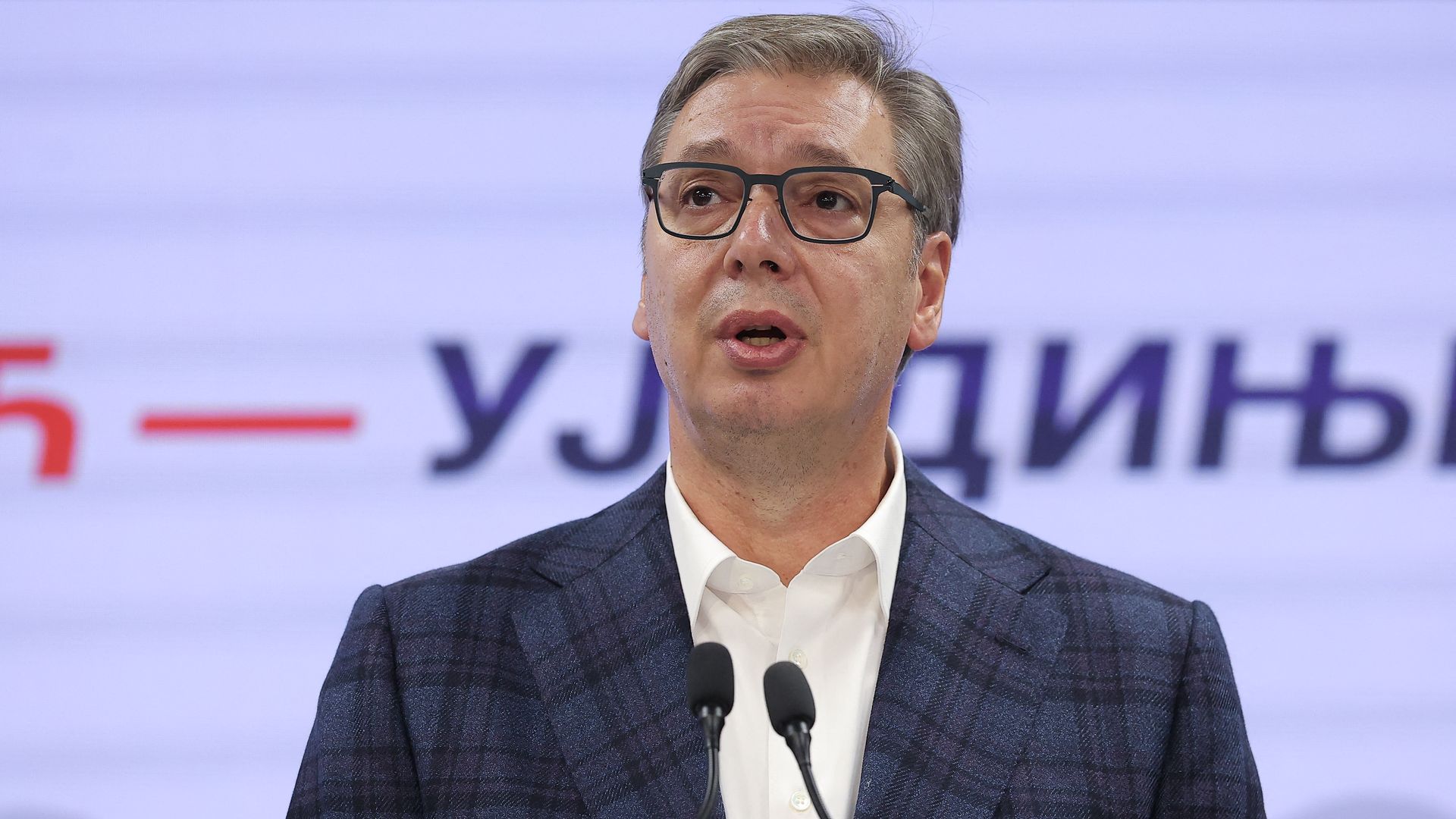 A middle-aged man with gray hair and glasses speaks at a podium, wearing a dark checkered suit and white shirt, with microphones in front and a blurred purple backdrop behind.