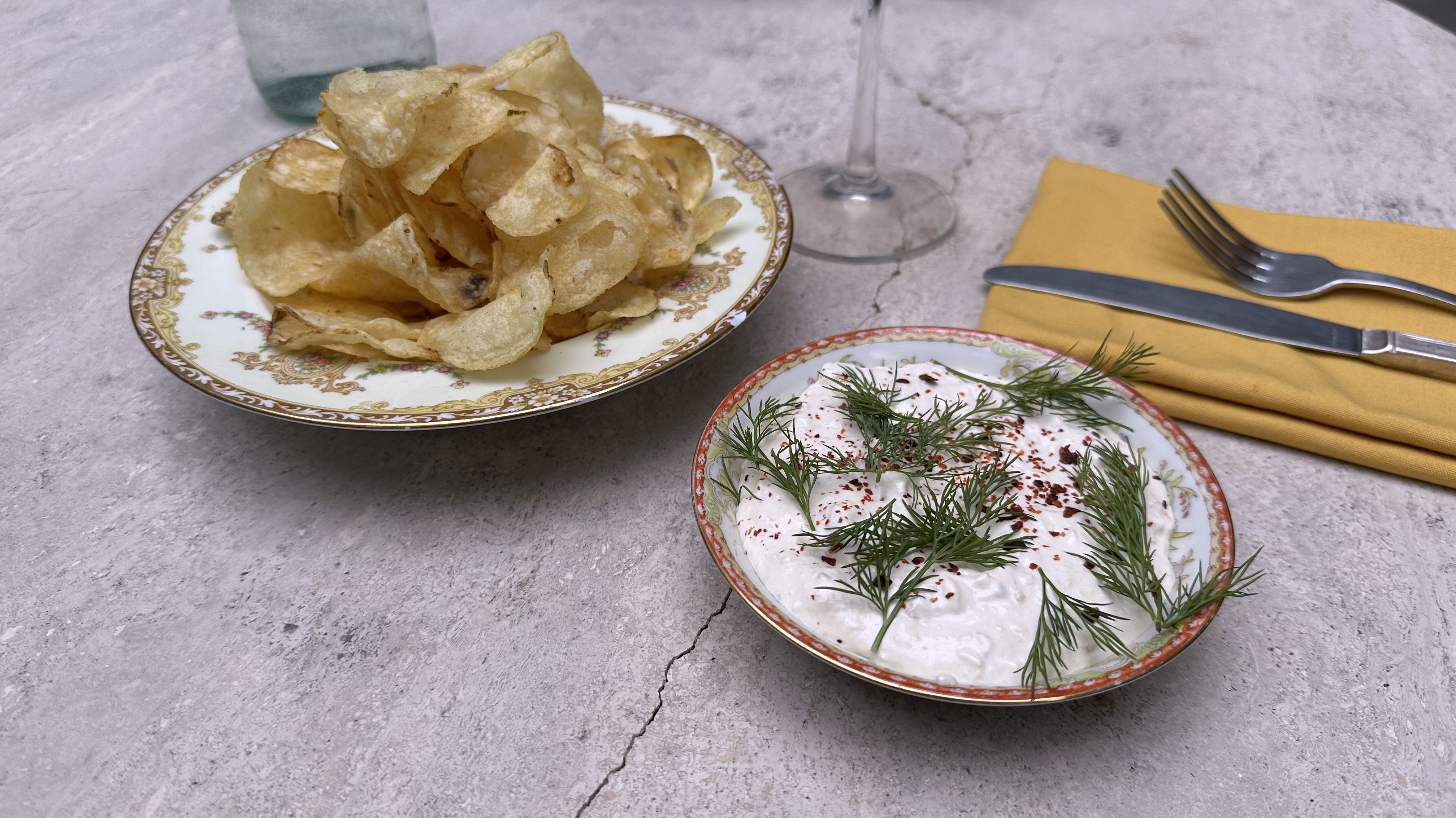 A marble table is set with a plate with potato chips, a small dish with a creamy dip topped with herbs, and a fork and knife on a yellow napkin.