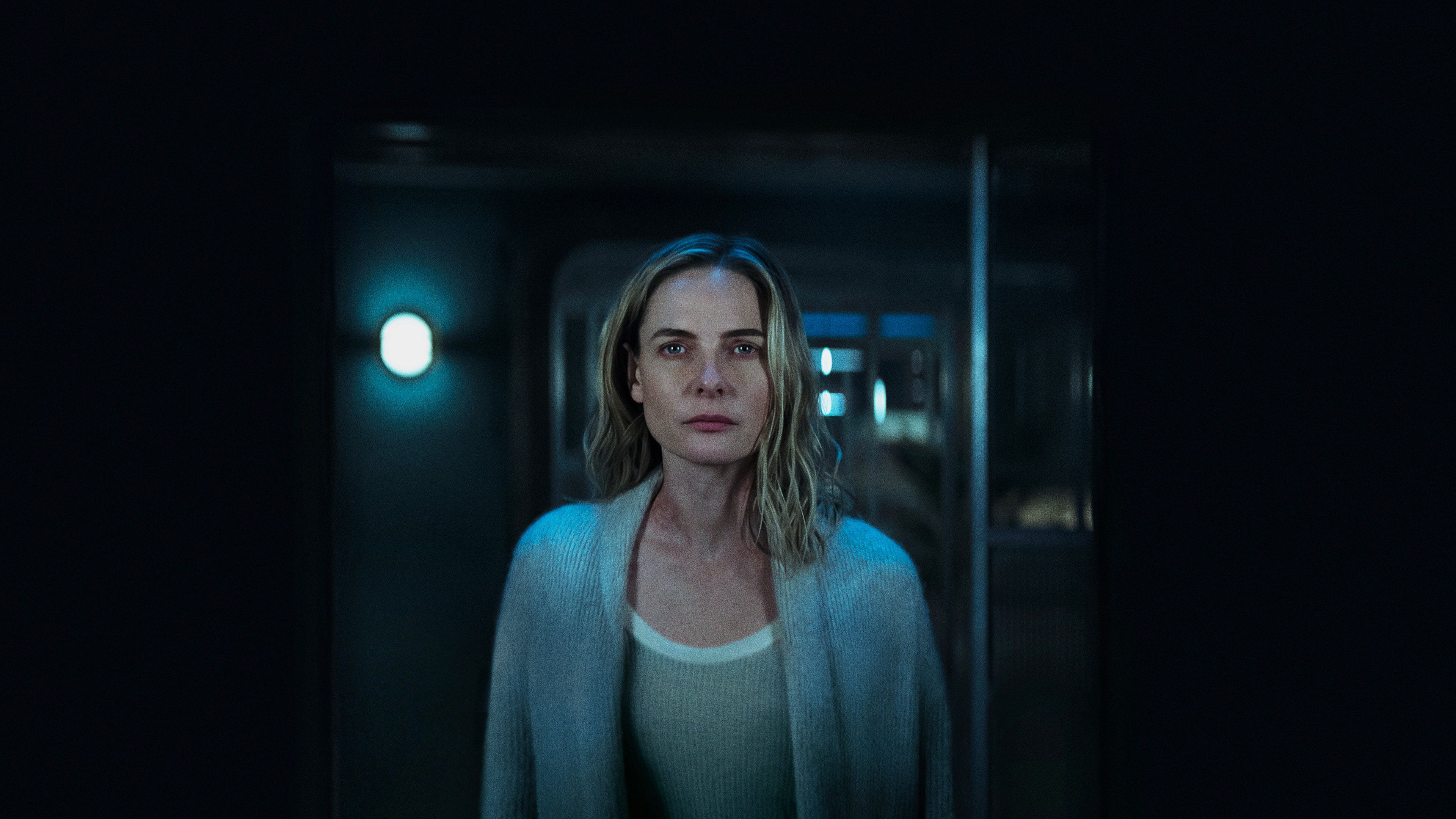 Rebecca Ferguson, with shoulder-length blonde hair, stands in a dim, blue-lit corridor, wearing a light cardigan over a white top, looking straight ahead with a neutral expression.