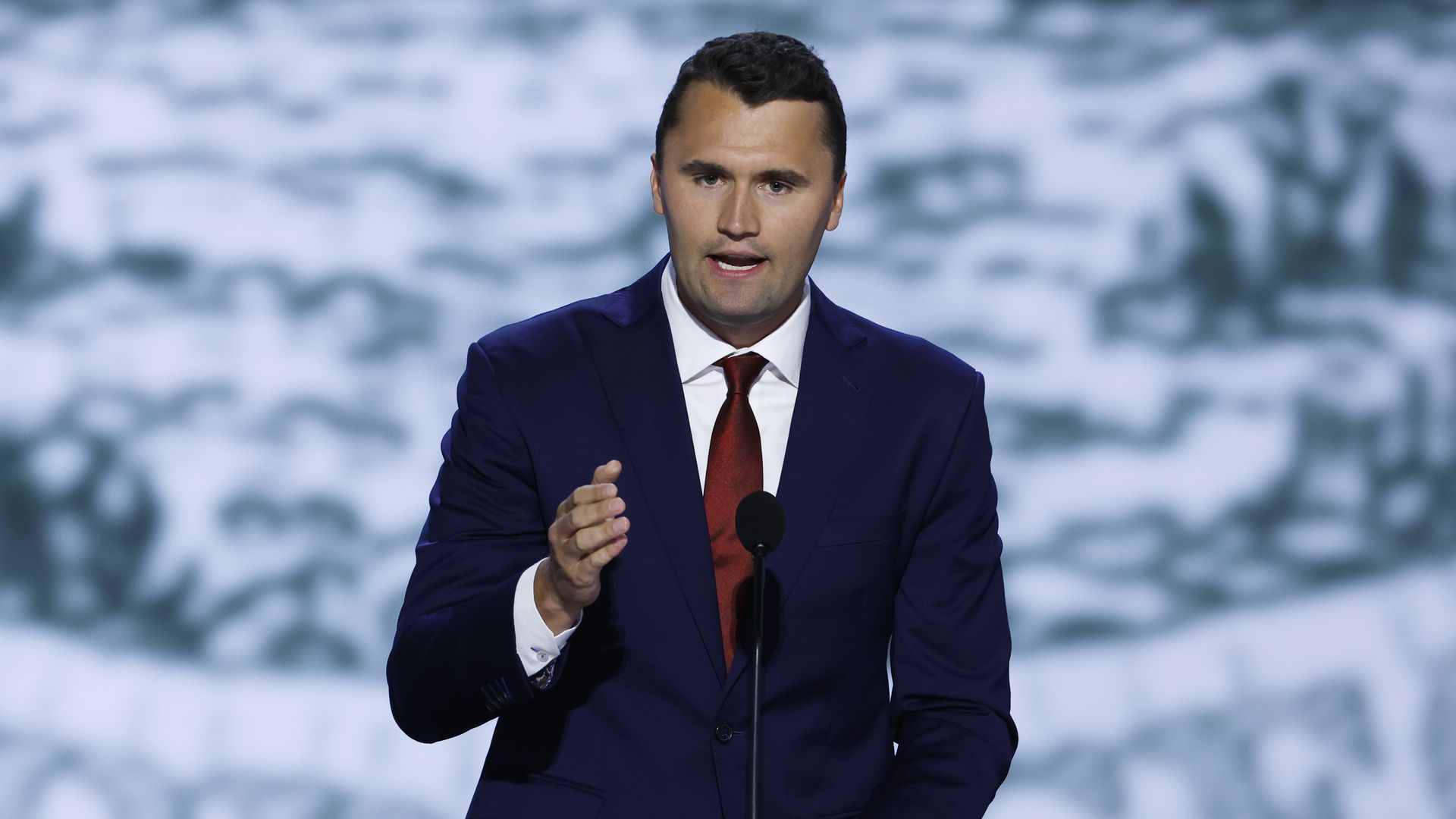 Charlie Kirk, wearing a blue suit and red tie, gestures with his hand while speaking on stage.