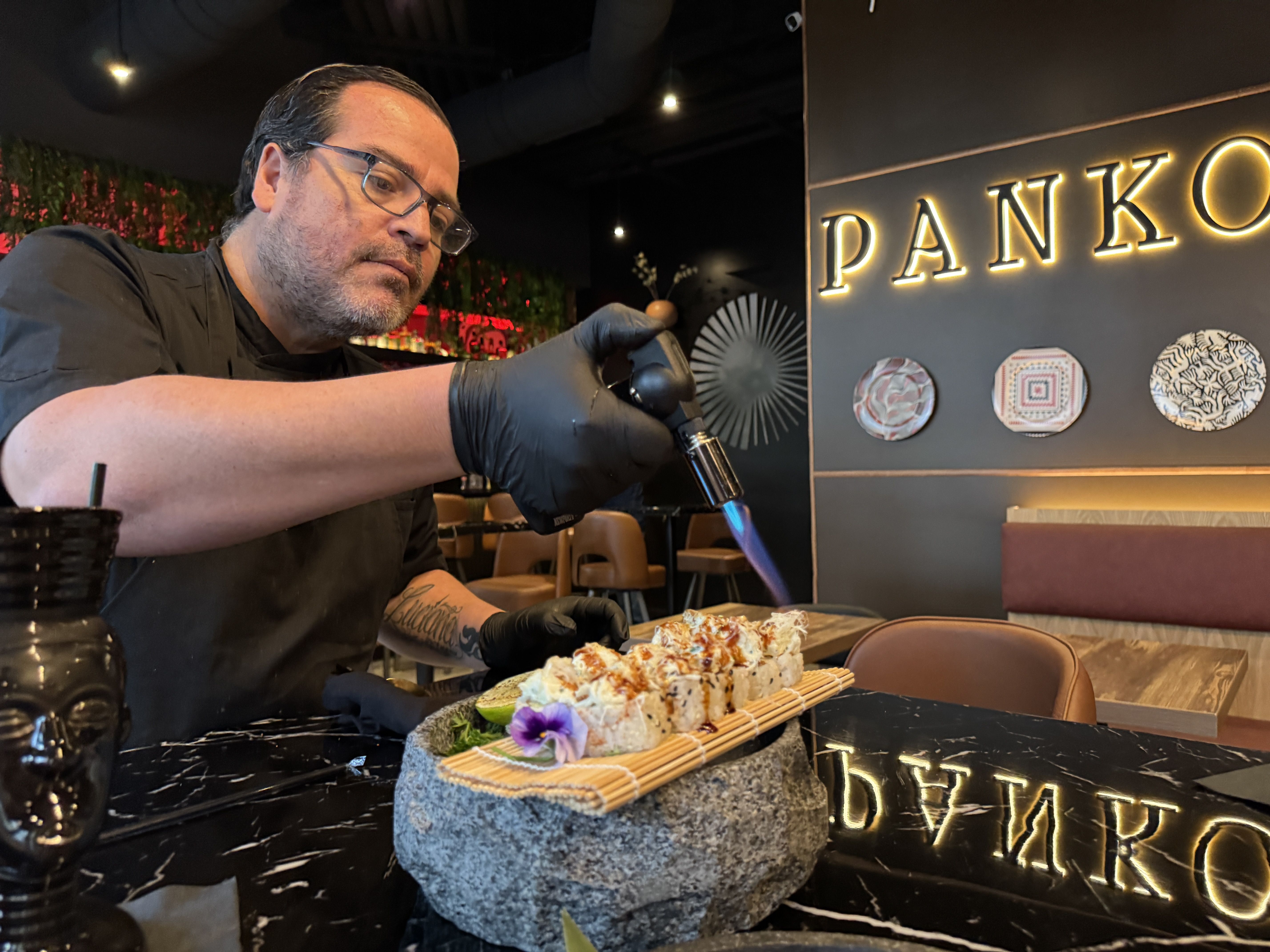 Chef with glasses and gloves using a blowtorch on sushi rolls on a stone plate at a restaurant with illuminated "PANKO" sign in the background and patterned wall decor.