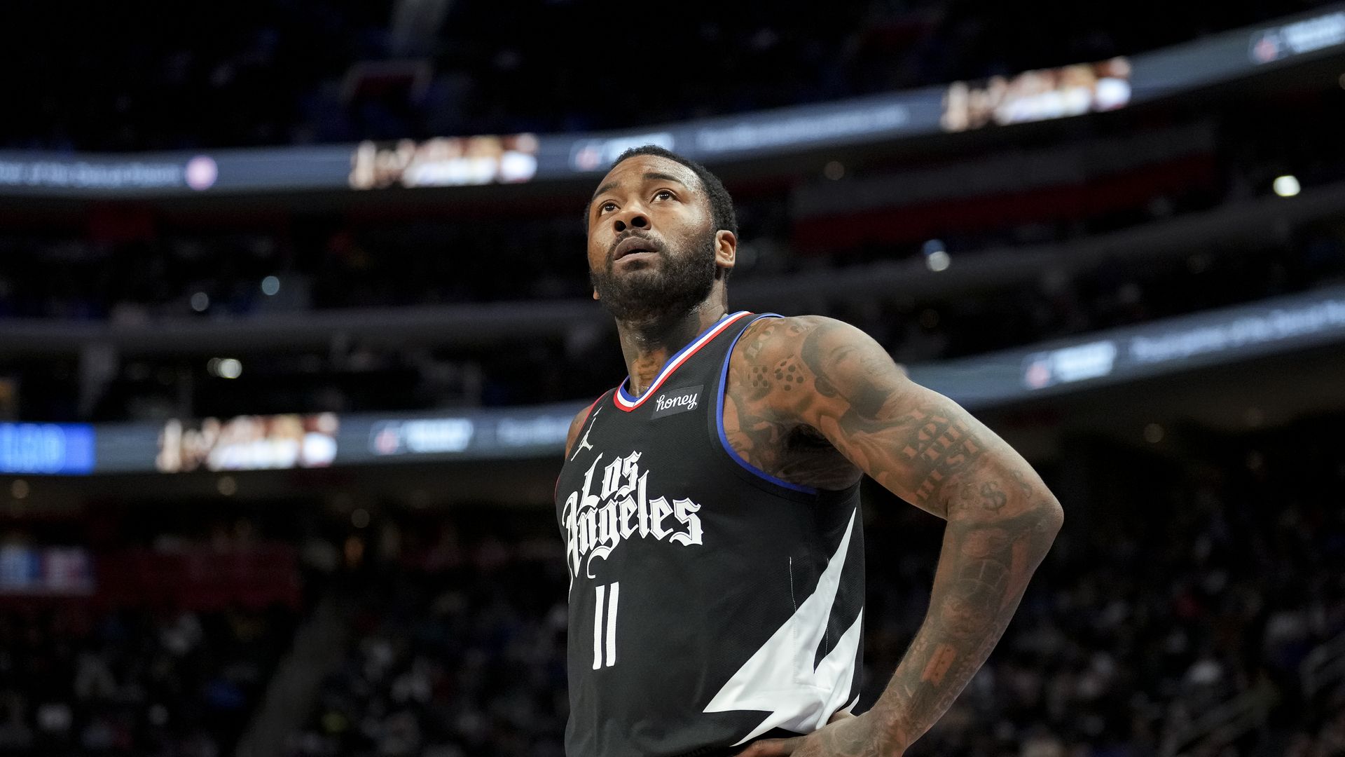 Basketball player wearing black Los Angeles jersey number 11 stands on court with hands on hips, looking upwards during a game in a crowded arena.