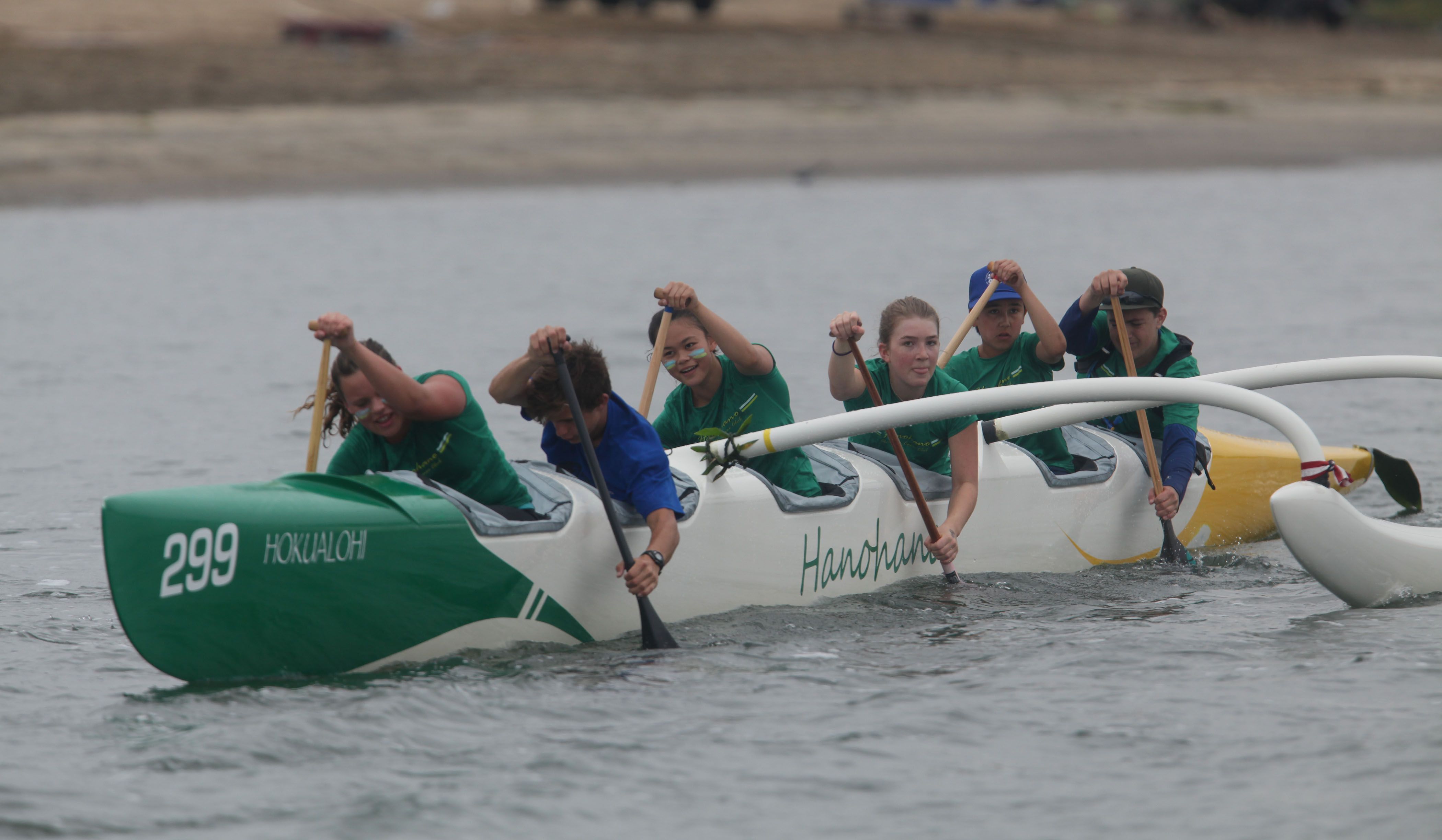 Dragon boat team in green shirts paddles in unison in a white-and-green boat marked 299, using wooden oars on calm water with a distant shoreline.