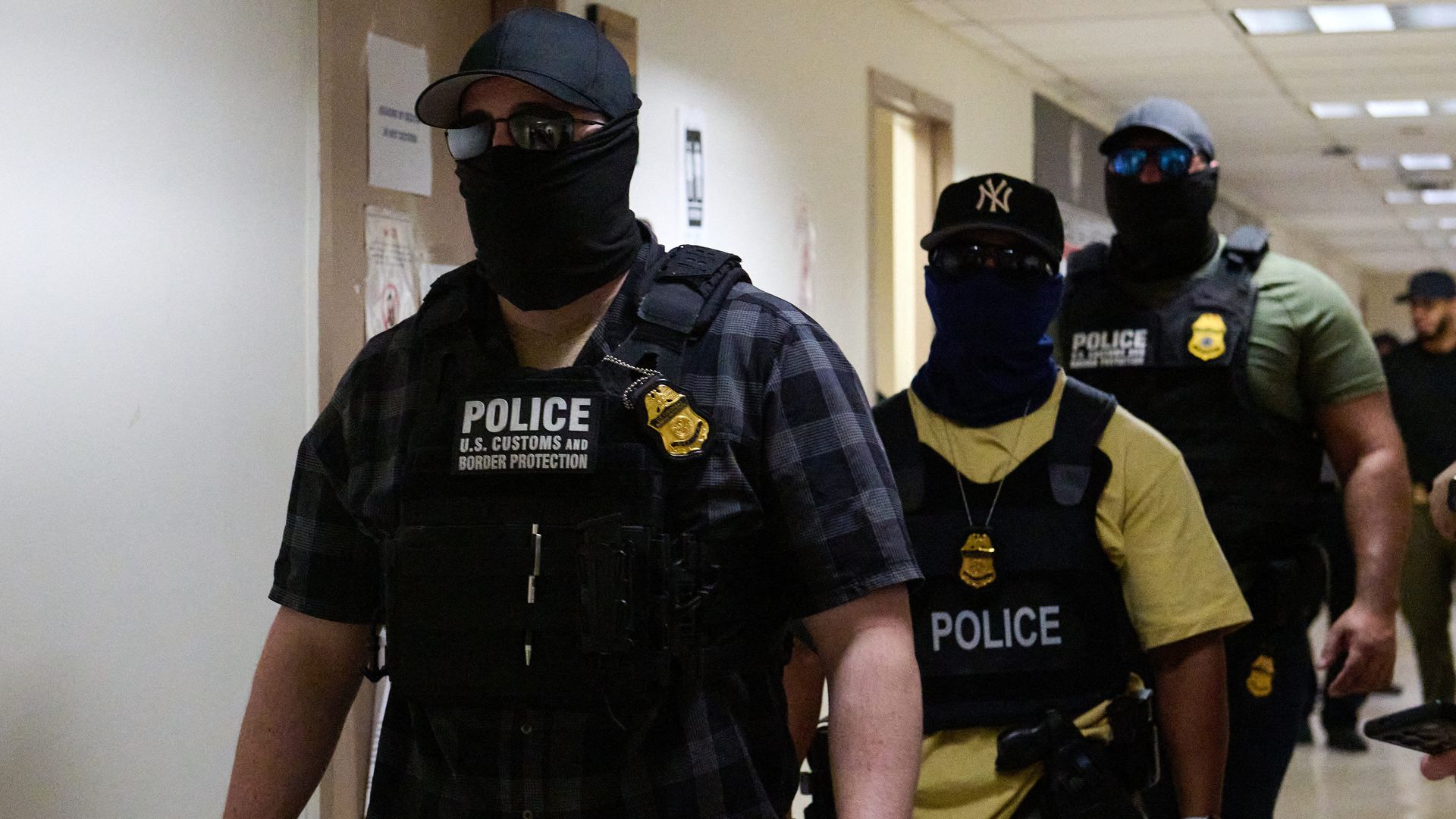 Three US Customs and Border Protection officers wearing black masks, sunglasses, police vests, and badges walk down a hallway with beige walls and fluorescent lighting.