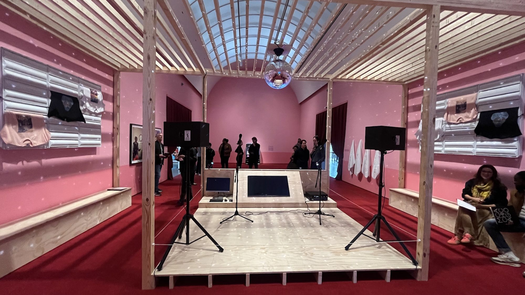 A pink exhibition room with a wooden stage, a suspended disco ball, and two microphone stands. Large speakers flank the stage as visitors mingle in the background.