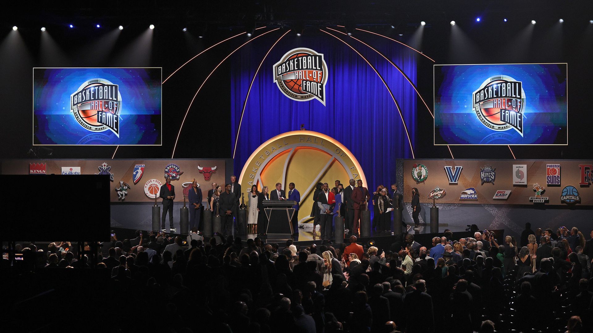 A stage surrounded by guests, filled with NBA stars and highlighted with big screen TVs showing the basketball hall of fame logo. 