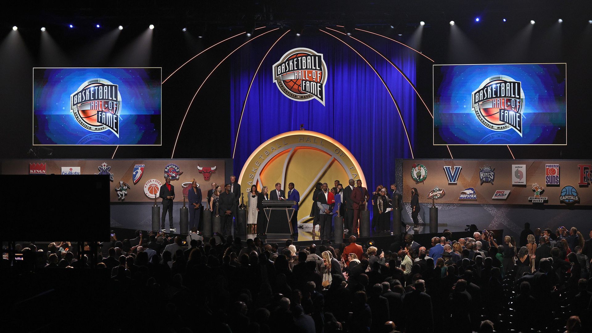 A stage surrounded by guests, filled with NBA stars and highlighted with big screen TVs showing the basketball hall of fame logo.