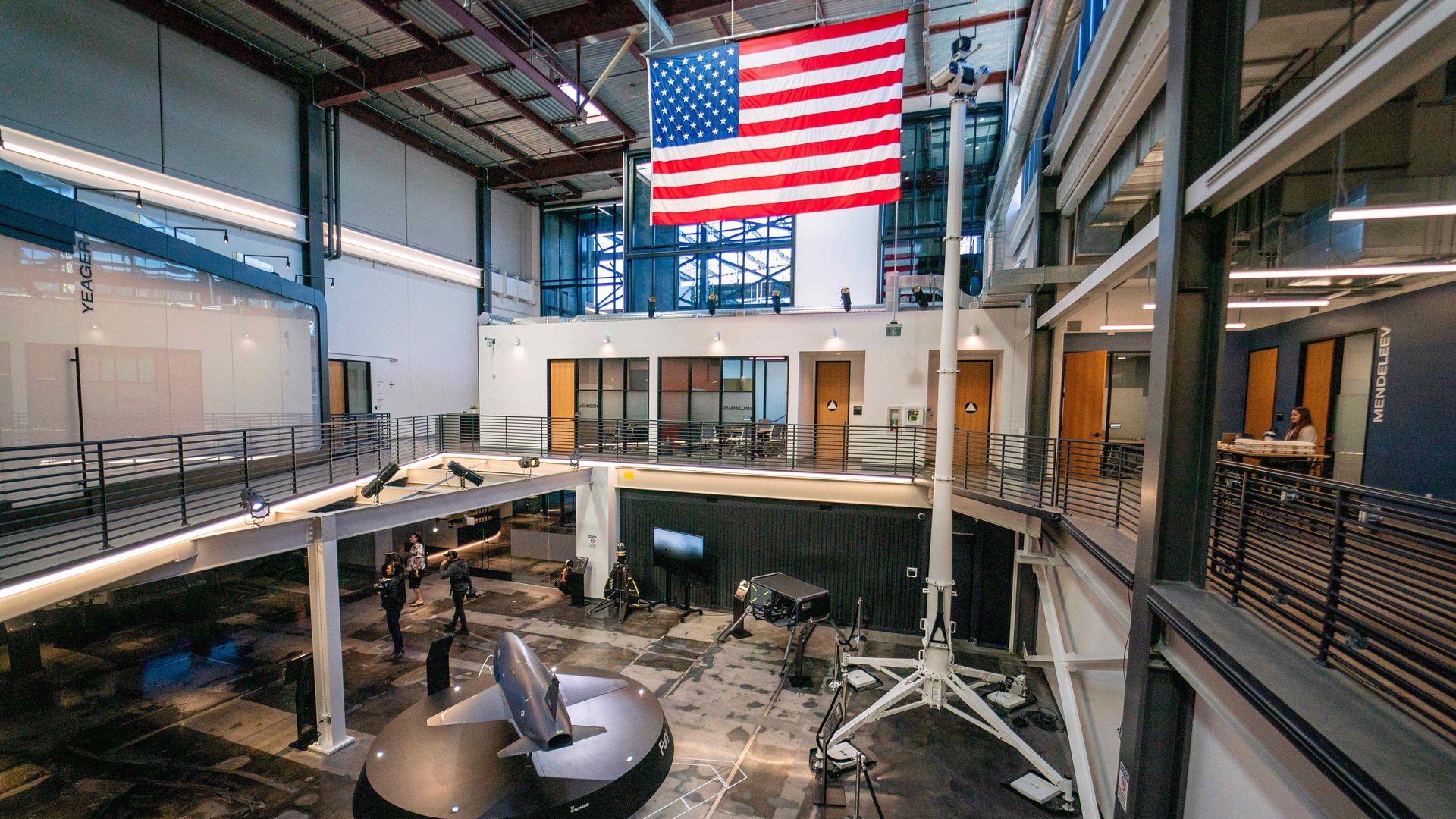 A large entrance to a defense-contractor's headquarters. Models of weapons and drones are seen on the bottom floor. A large American flag hangs from the rafters. People mill about. It is well lit.
