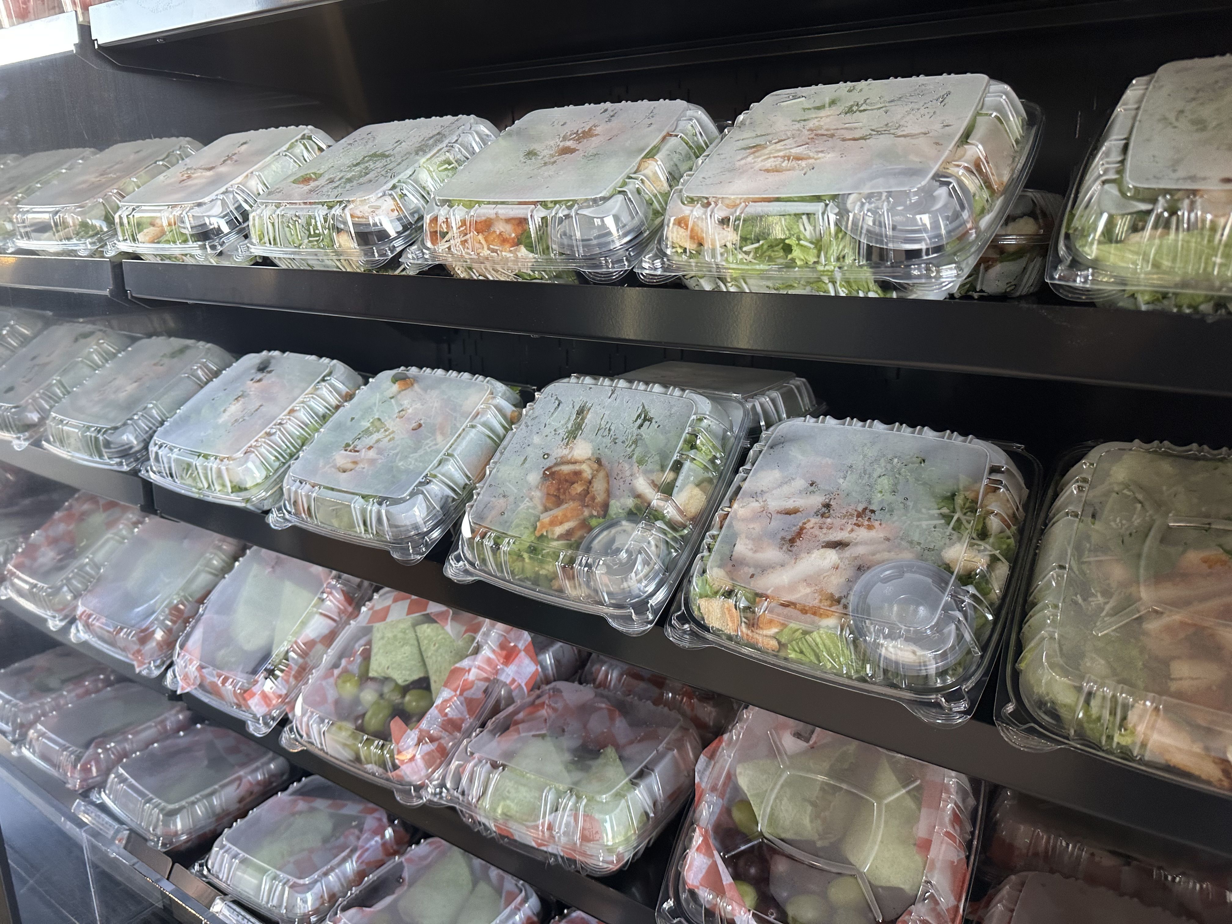 Rows of clear plastic takeout containers on metal grocery-store shelves, filled with salads and prepared meals; greens, chicken, and vegetables visible through the lids.