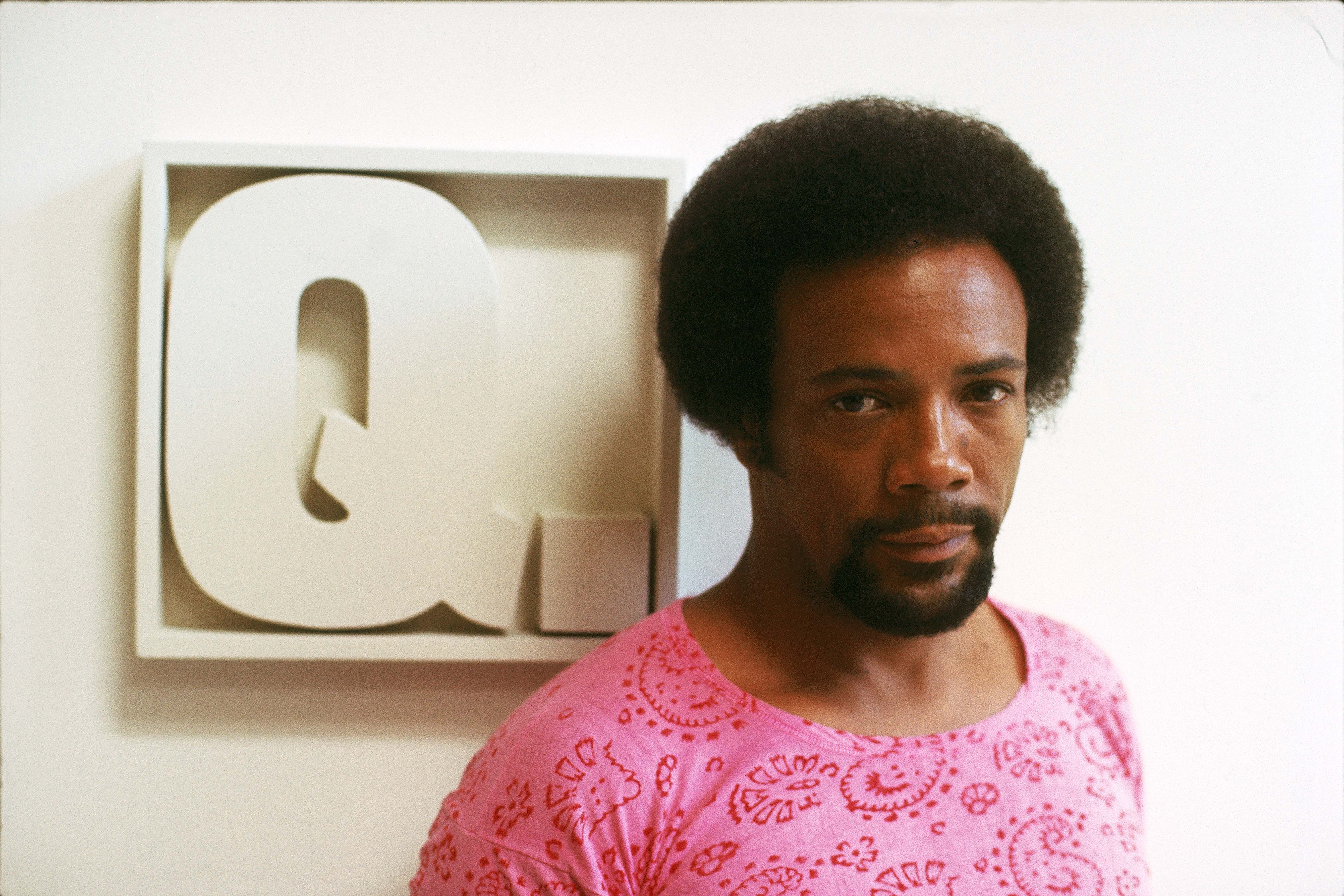 A posed photo of Quincy Jones in a pink shirt in front of a shadow box on a wall.
