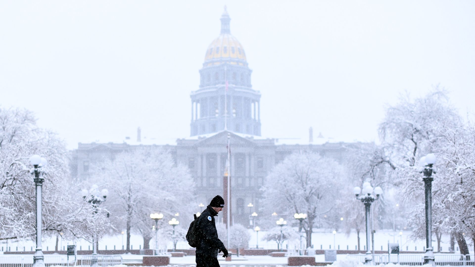A man walks in a snowstorm as a massive building is dusted by snow in the background. 
