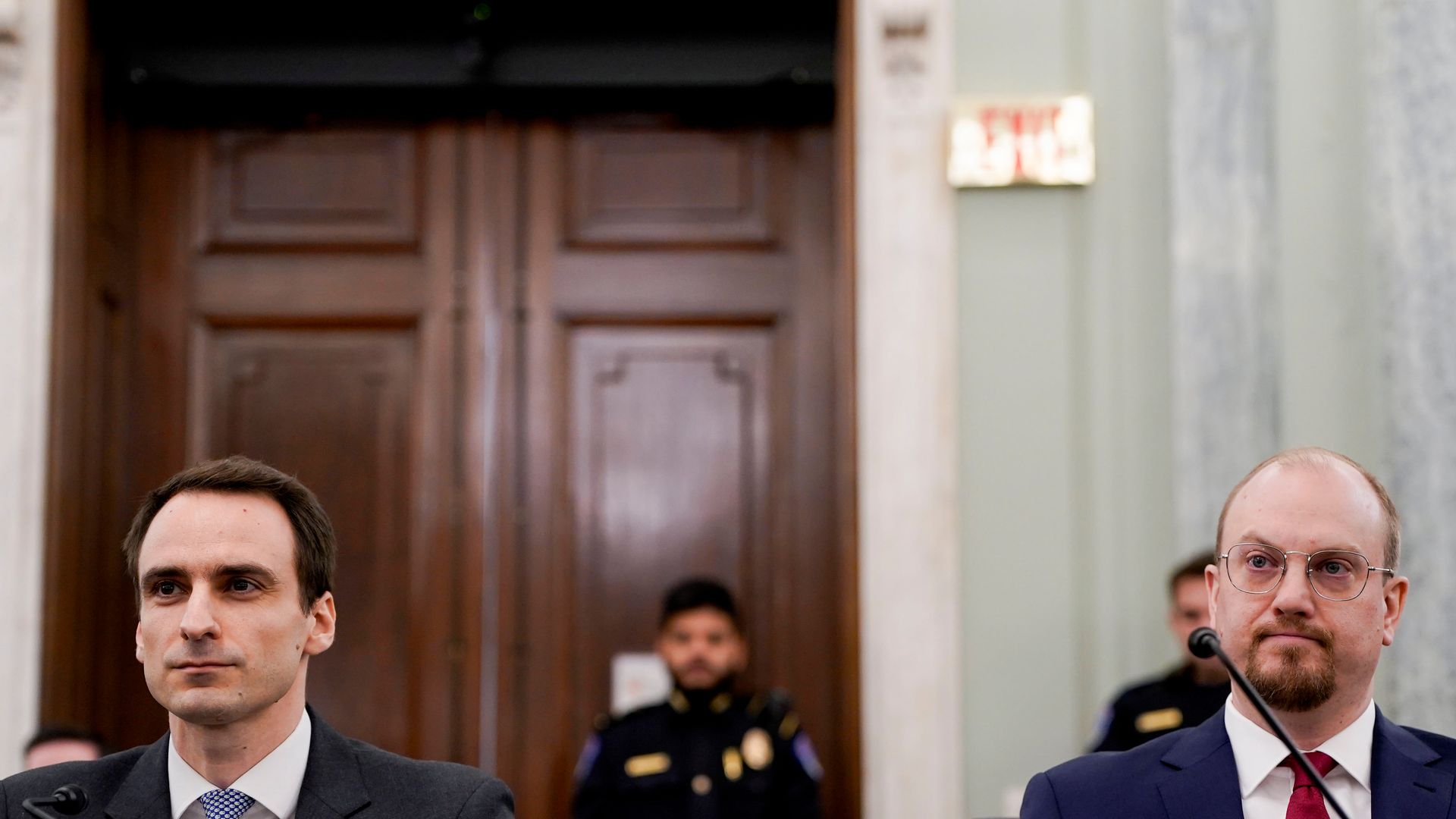 Two people in suits and ties sit in front of microphones