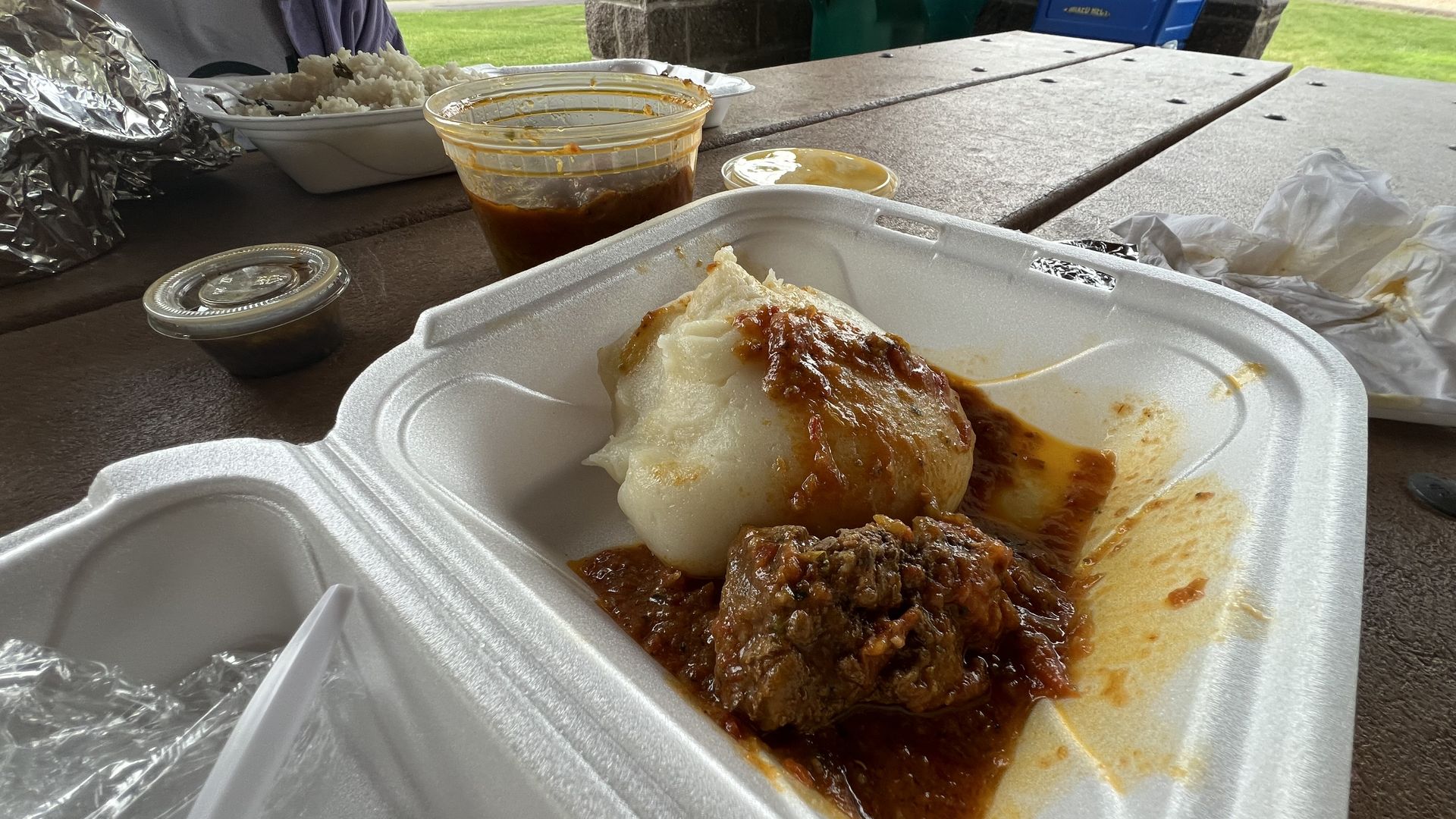 Foam takeout container with a serving of white soft food (fufu, a west African dish) topped with red sauce and a piece of meat on a picnic table, with sauce cups and a person eating in the background.