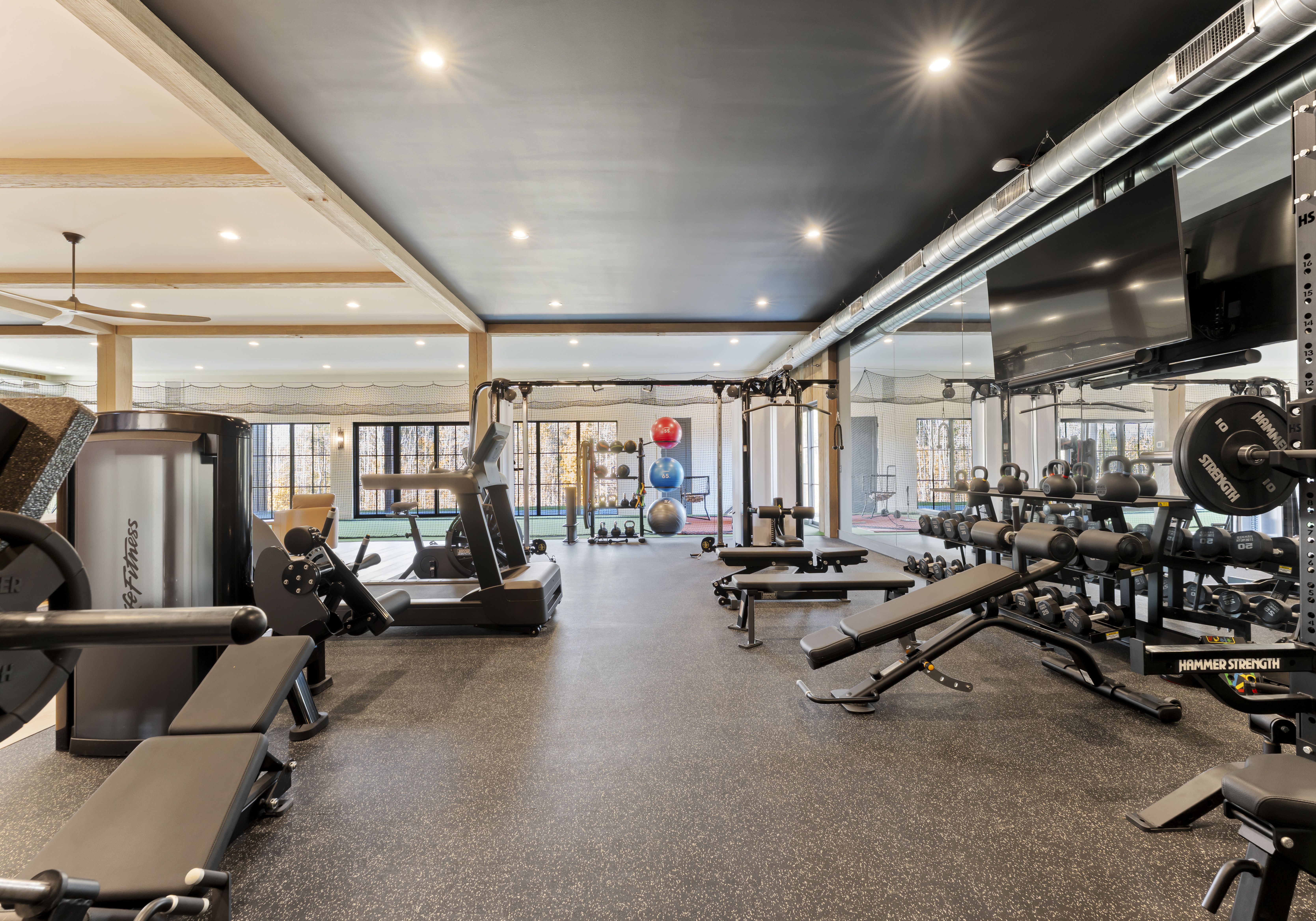 Wide gym interior with treadmills, benches, and dumbbells along both walls. Colorful medicine balls near back windows; black ceiling, exposed ducts, bright overhead lights.