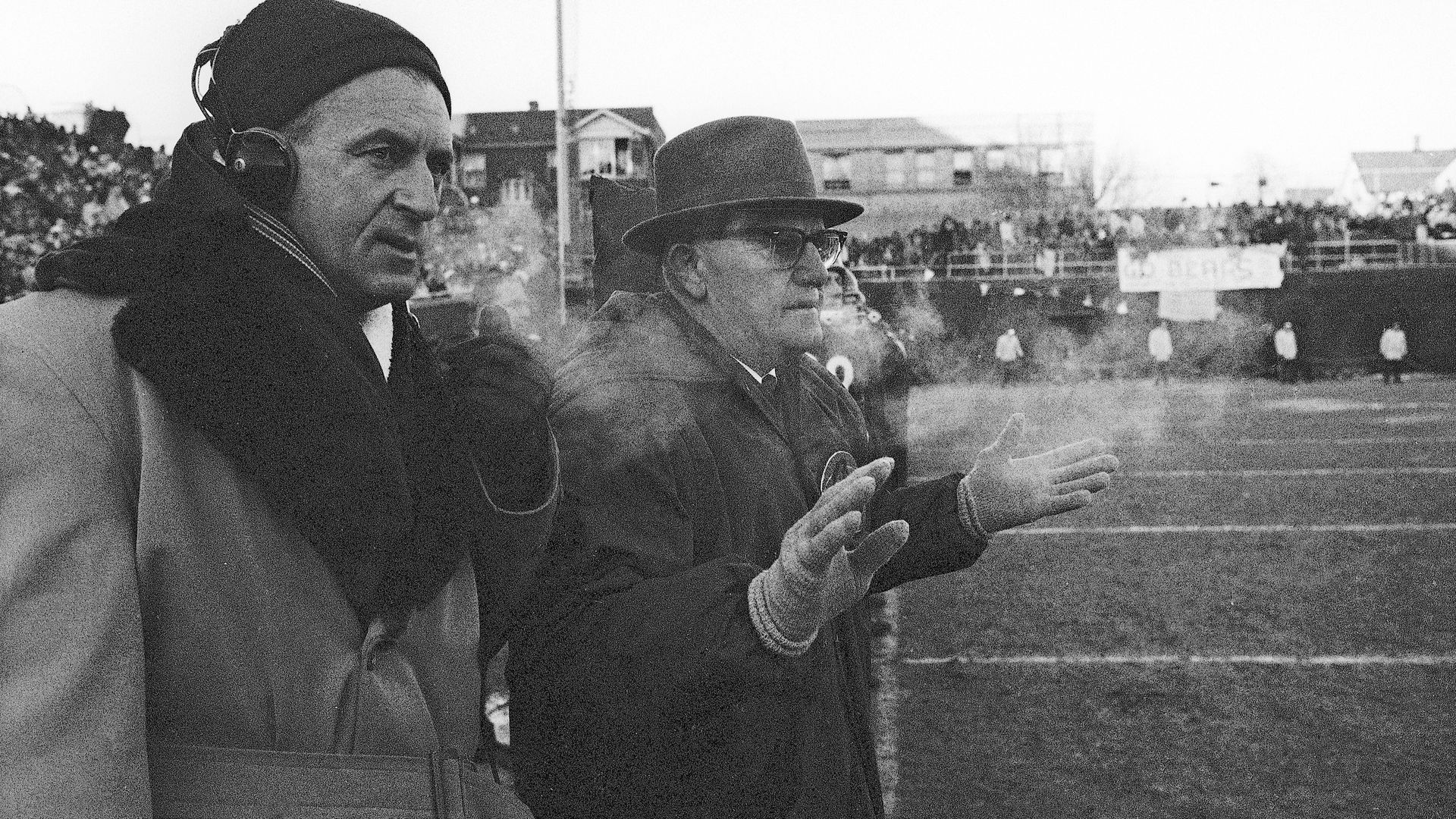 Former Bears coach George Halas on the sidelines of the field next to a man wearing headphones. 