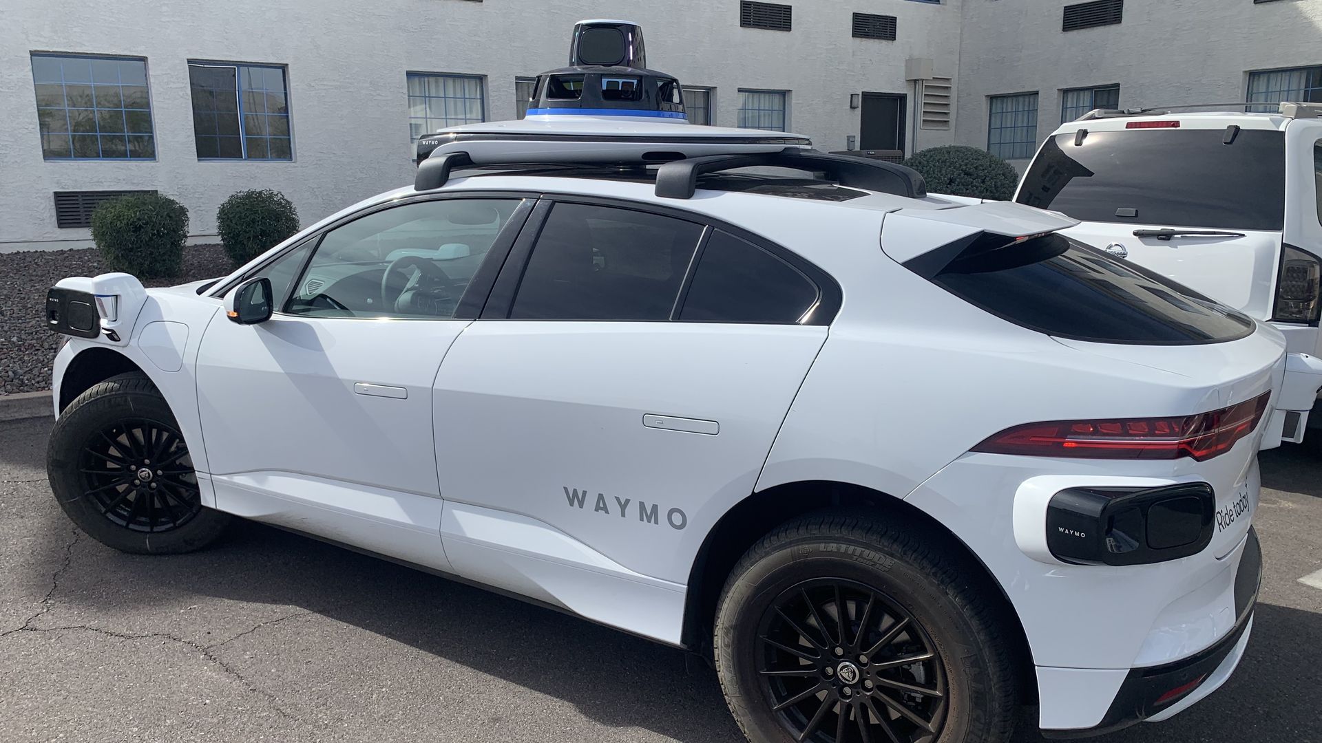 White Waymo self-driving car with black rims and sensors on front, back, and roof, parked in lot next to white building with blue windows.