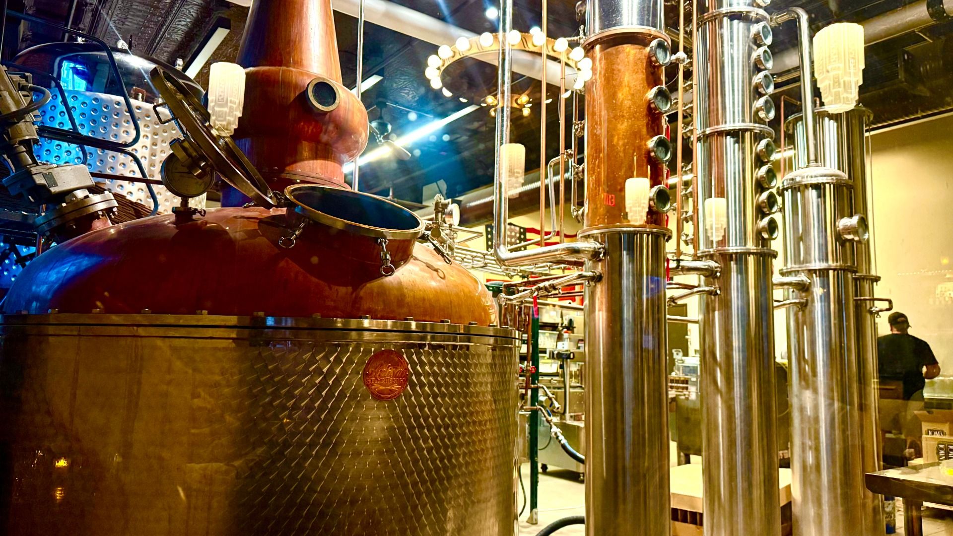 Inside a distillery, a large polished copper pot still with a domed top and connected stainless steel column stills. Bright lights, metallic pipes, and a person in the background.