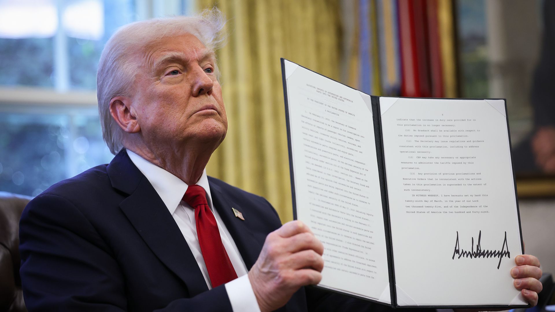 President Trump, seated in the Oval Office, holding up a signed executive order. 