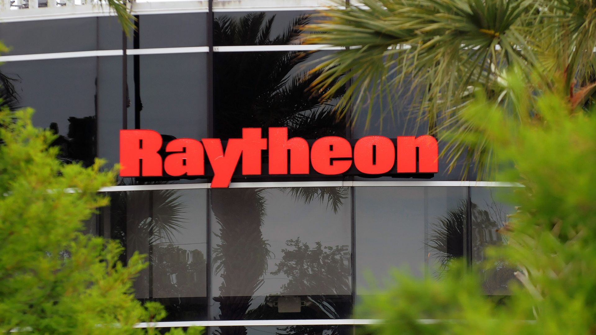 In this image, the Raytheon logo is seen on the side of a glass building, framed by trees.
