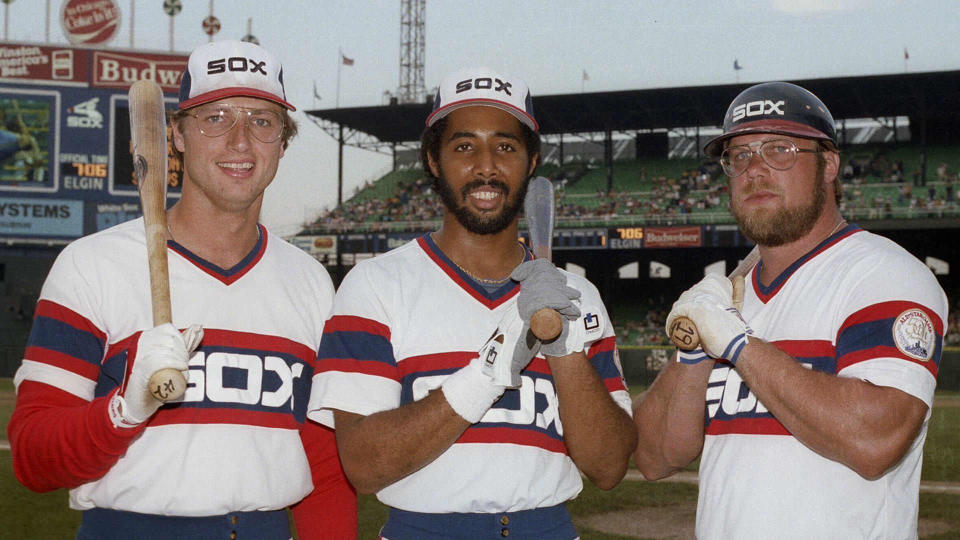 Three Chicago White Sox baseball players in white uniforms with red and blue stripes, holding bats, standing on a baseball field with stadium seats and lights in the background.