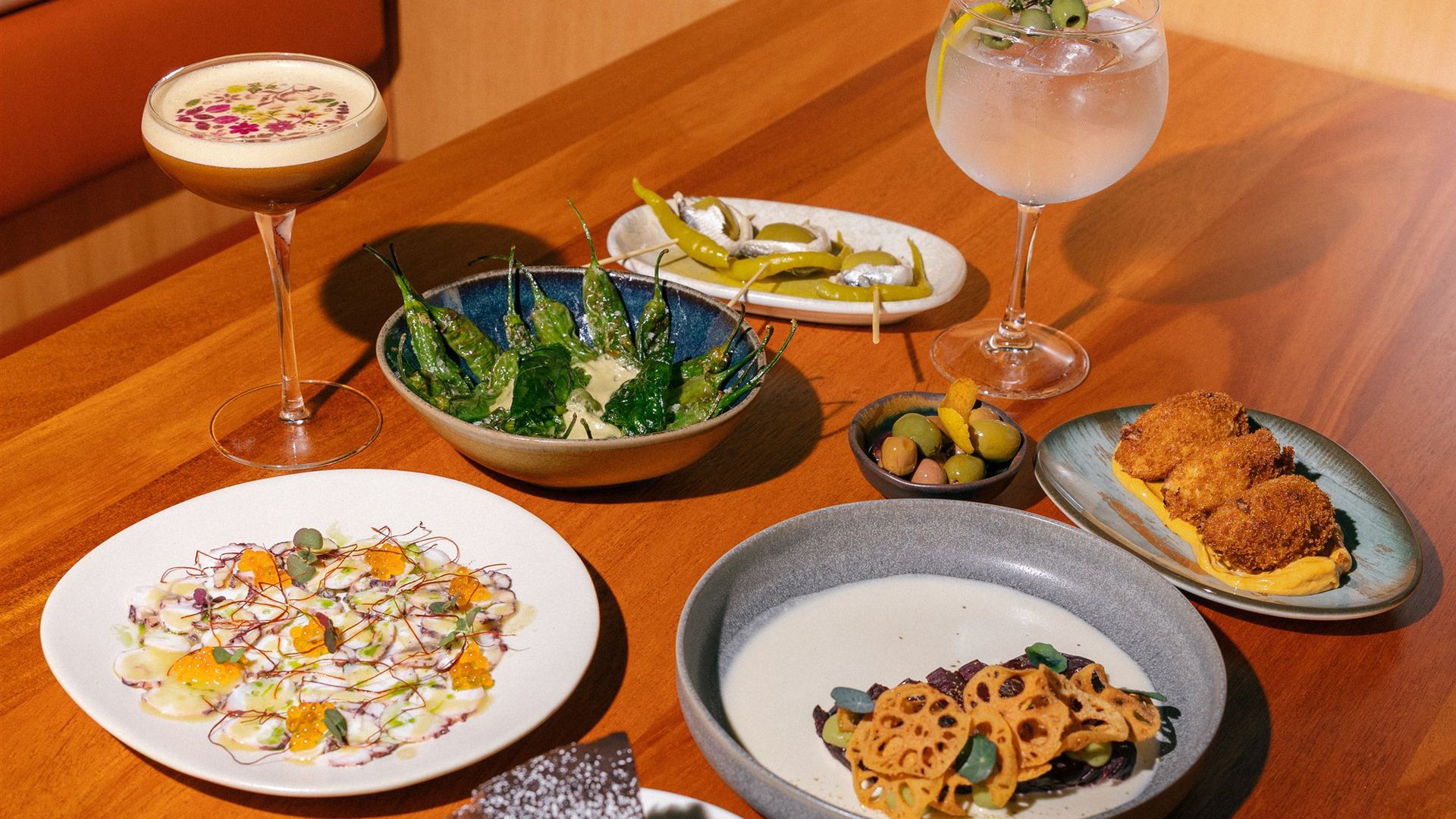 Assorted tapas on a table: blistered peppers with dip, olives, lotus-root chips, croquettes with yellow sauce; two cocktails, one foamy with flowers, the other clear with citrus.