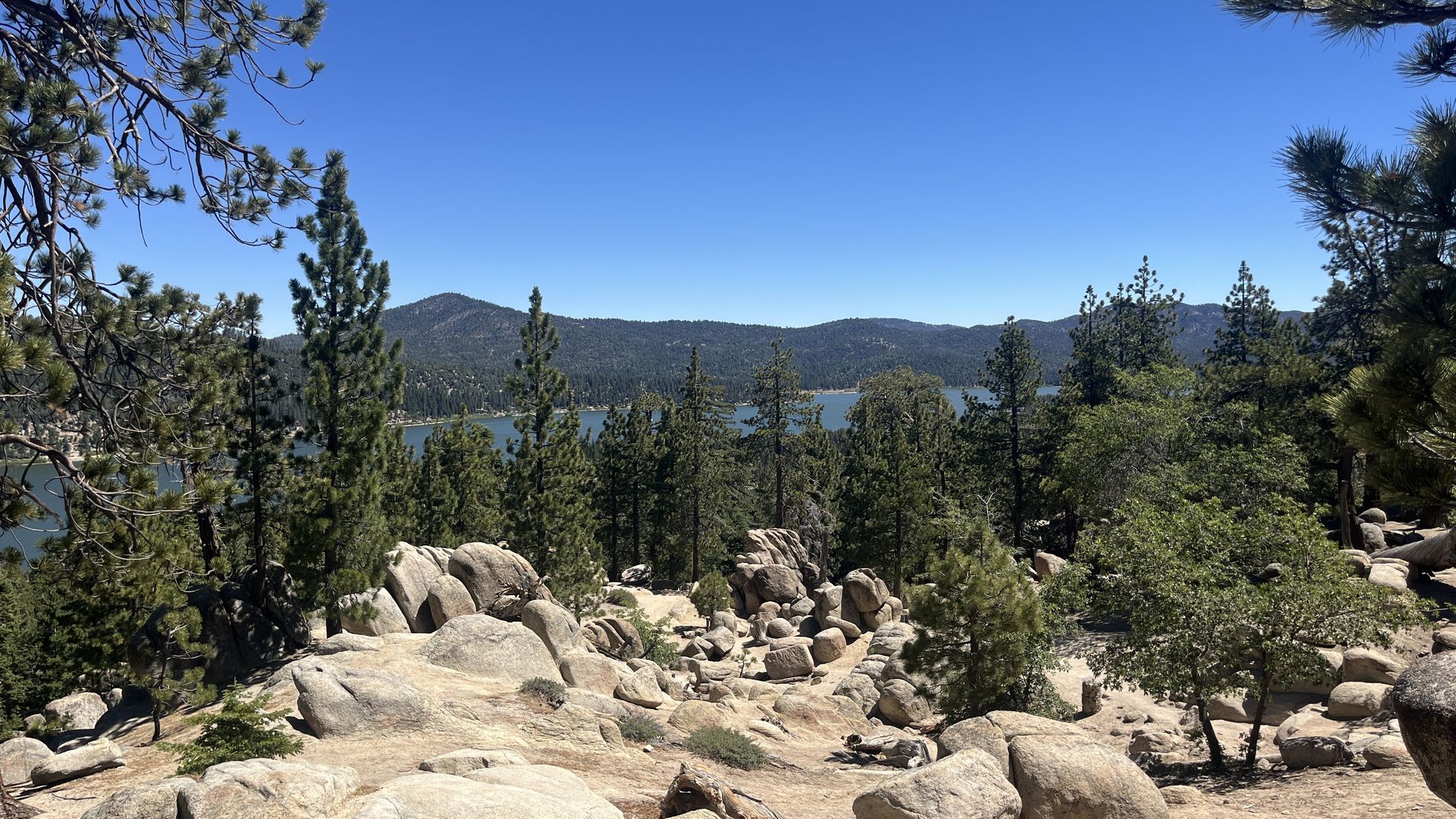 Panoramic view from a rocky overlook: large boulders in the foreground, tall pine trees, a blue lake bordered by forested hills, under a clear blue sky.