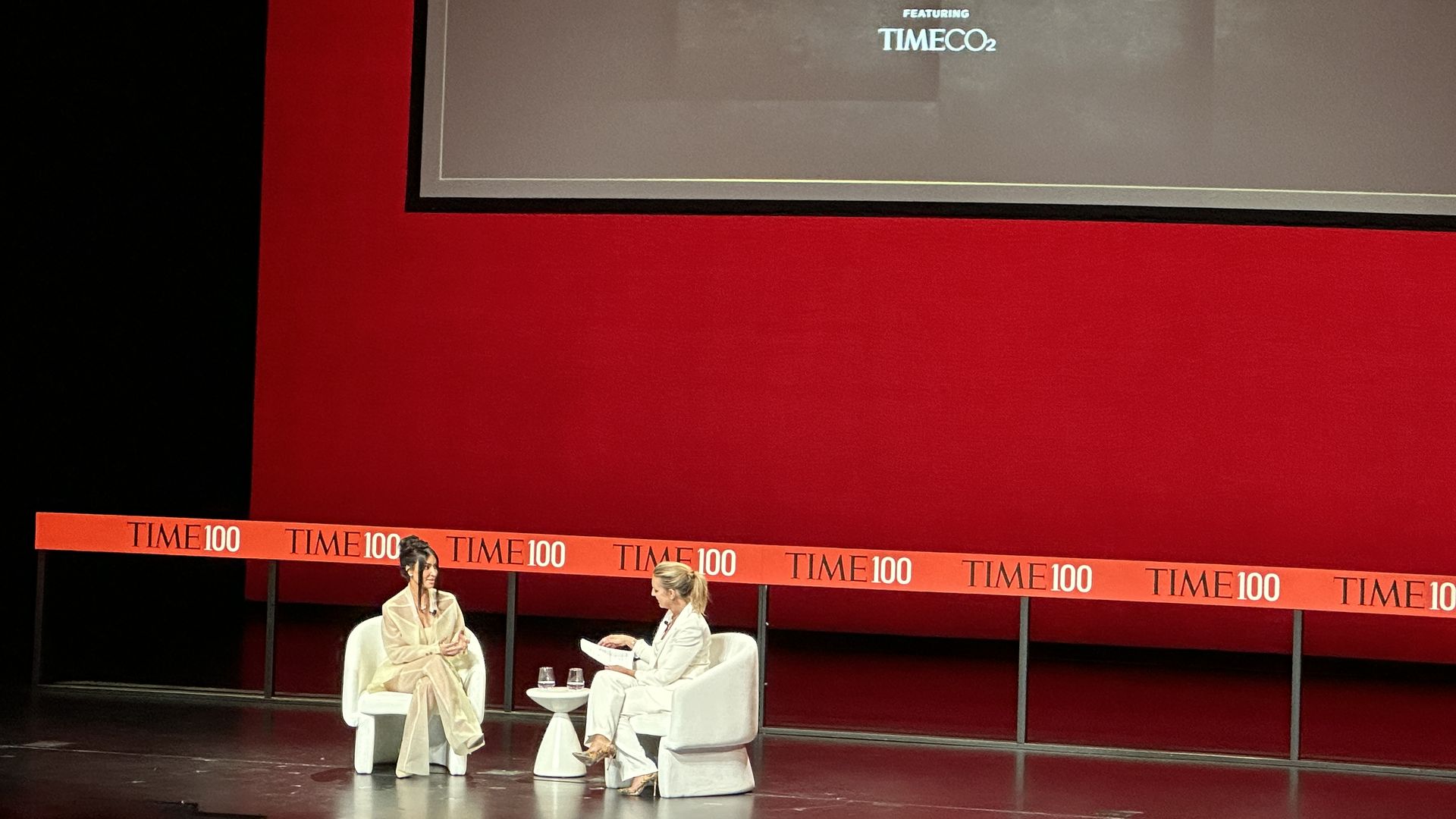 Kim Kardashian and Poppy Harlow both sitting in white chairs facing each other and talking in front of a red backdrop at the TIME100 Summit 
