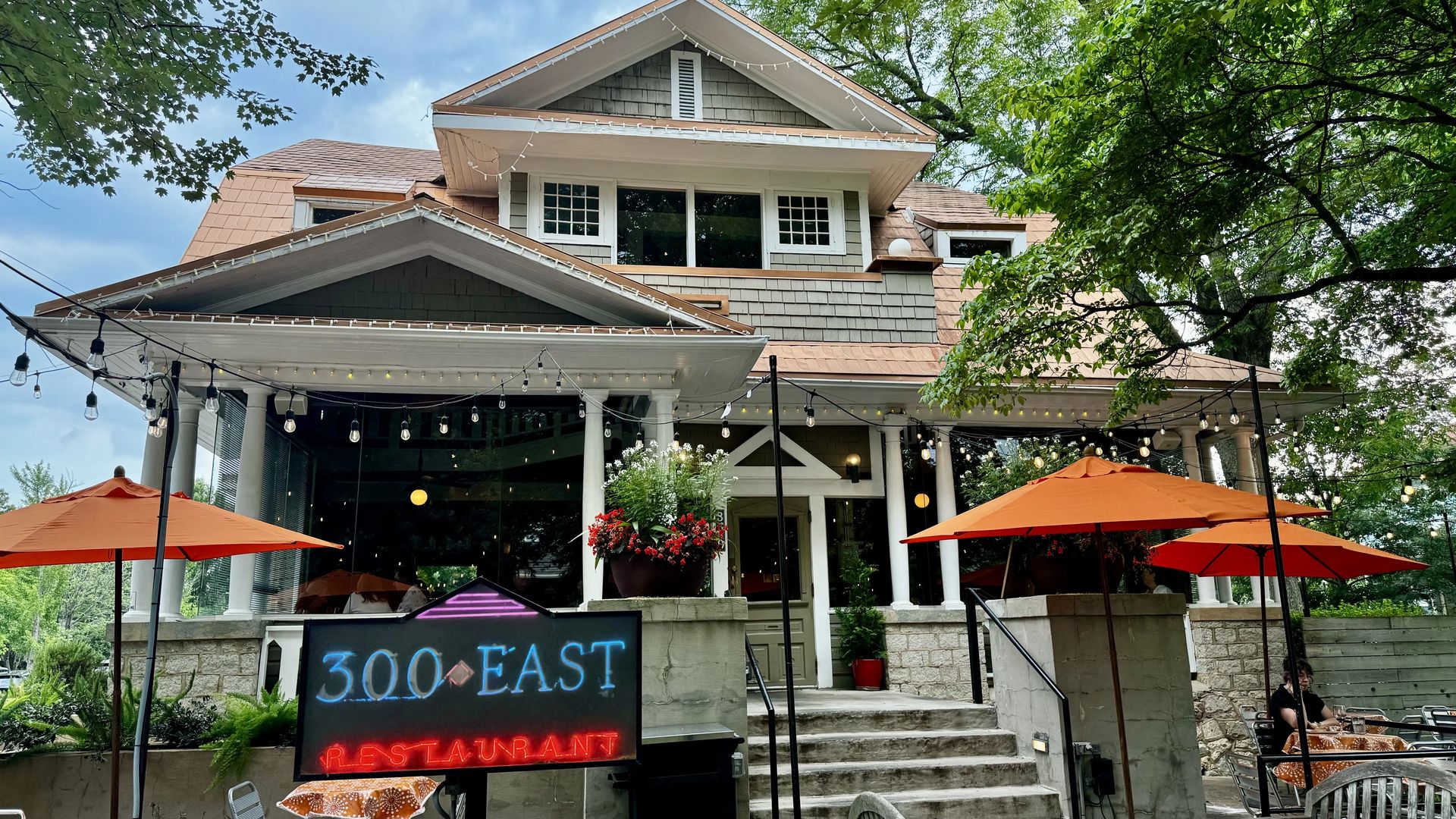 A charming, two-story house in a residential area converted into a restaurant, with a sign reading "300 East Restaurant". The house features a large porch with string lights, red flowers in planters, and outdoor seating under orange umbrellas. 