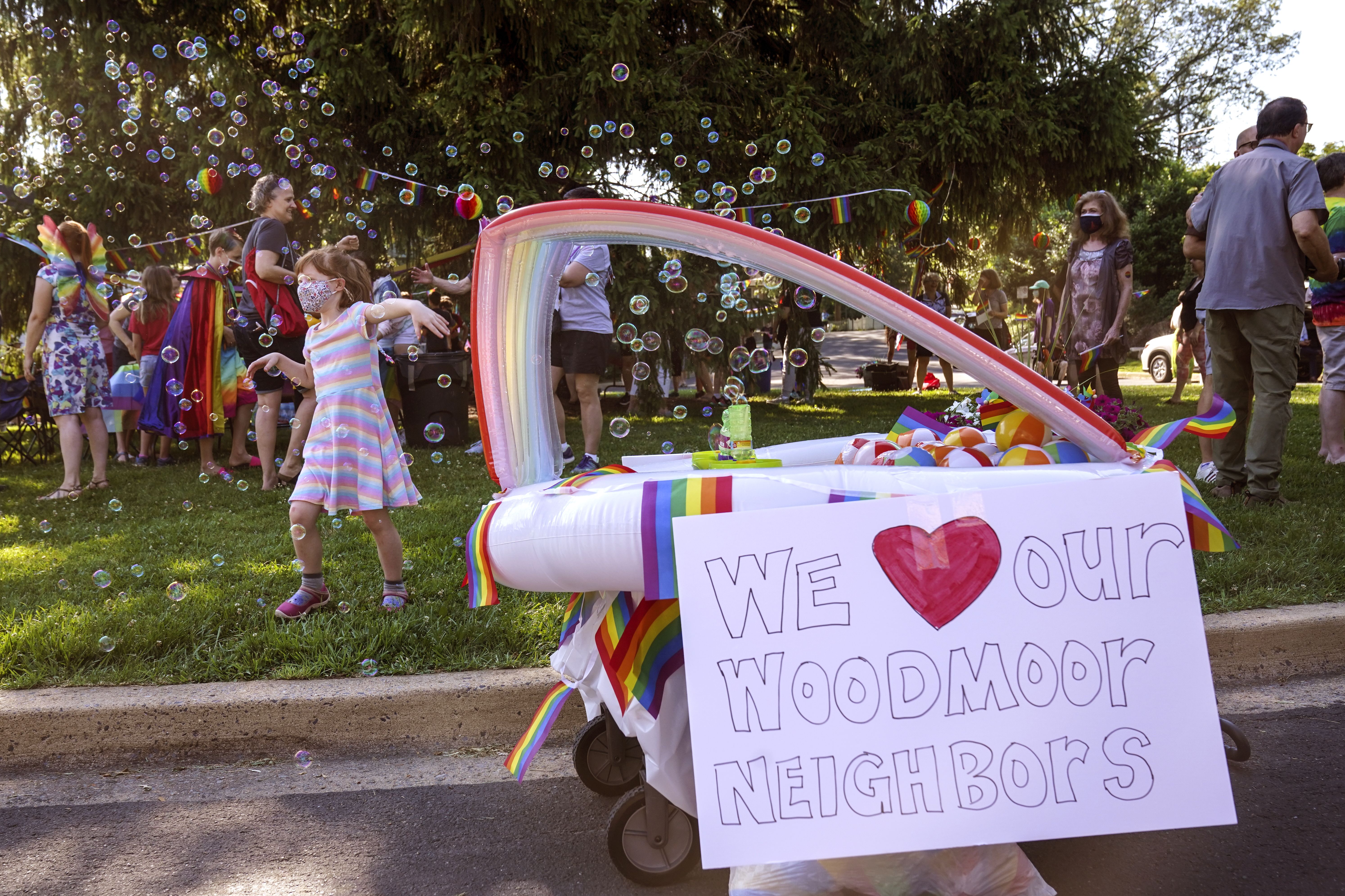 Photo of a bunch of people on the grass wearing a variety of rainbow colors, and a cart with a sign that says "We heart Woodmoor neighbors"